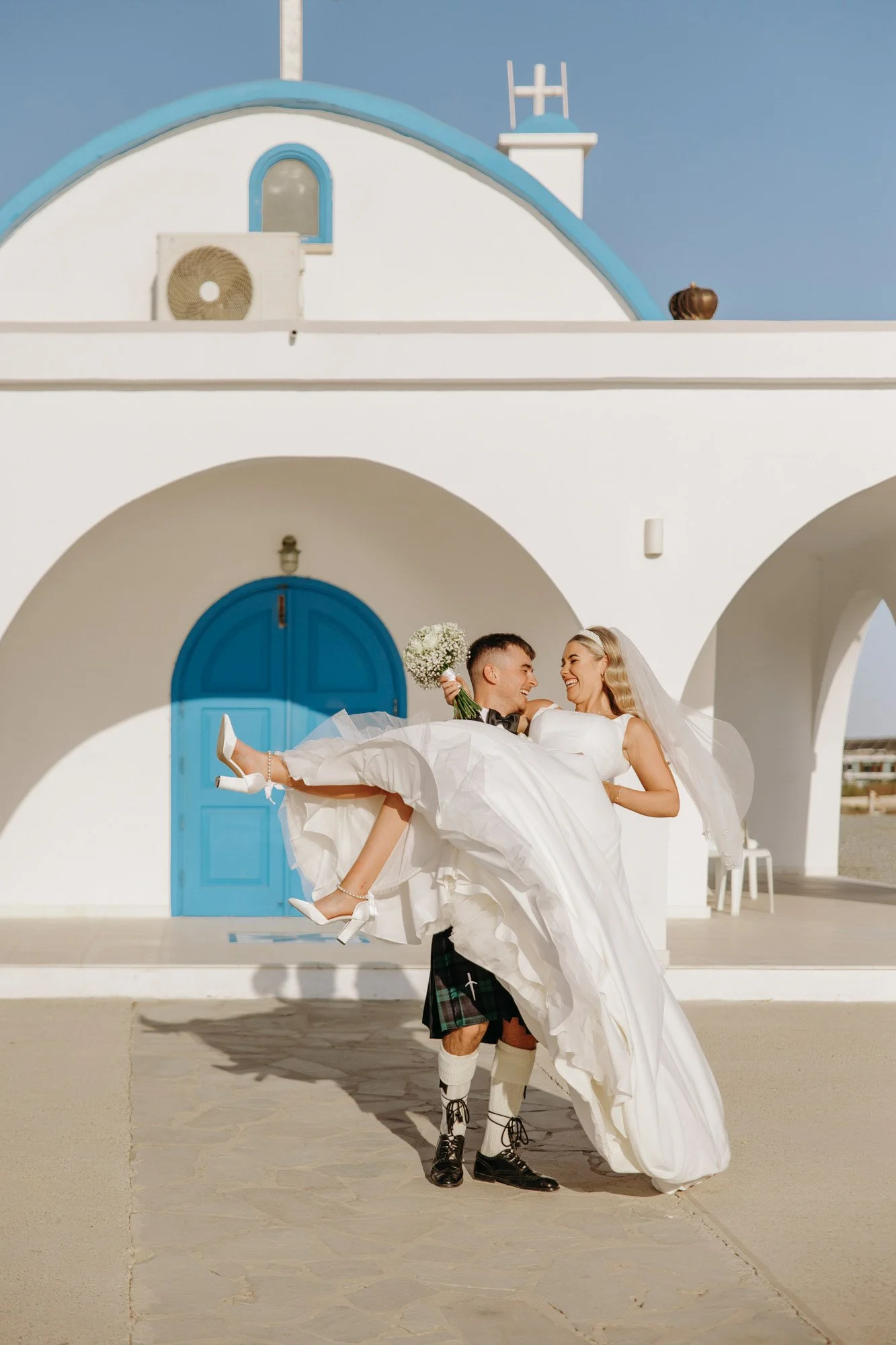 A bride and groom celebrating on a sunny day outside a white building with a blue door and accents. The groom is dressed in traditional Scottish attire, including a kilt and black shoes, and is carrying the bride who is wearing a white wedding gown a