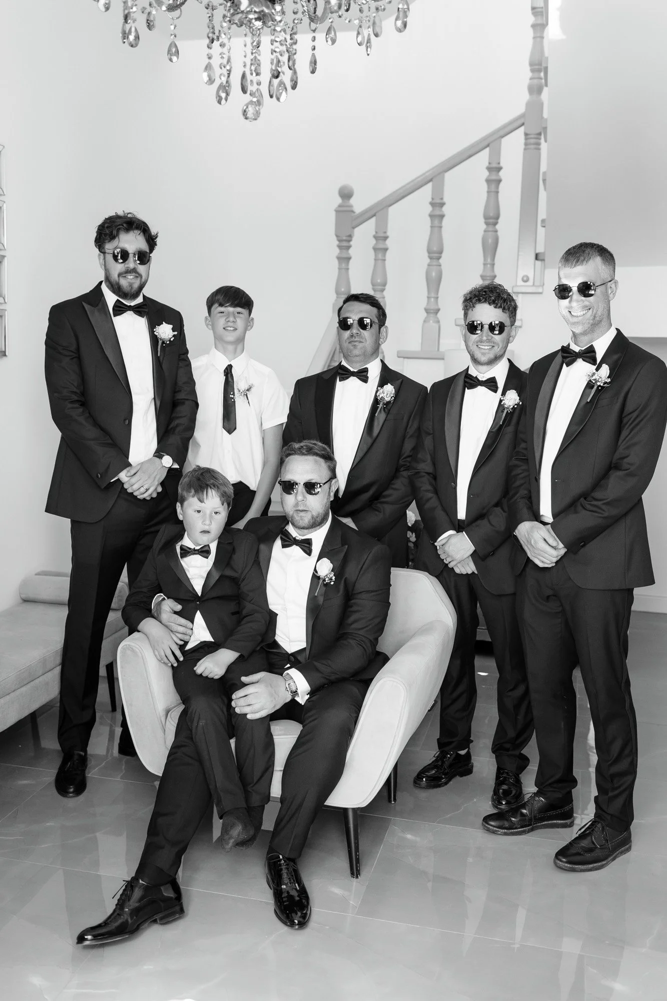 A group of eight men and boys wearing formal tuxedos and sunglasses, posing together in a living room with a chandelier overhead and a staircase in the background.