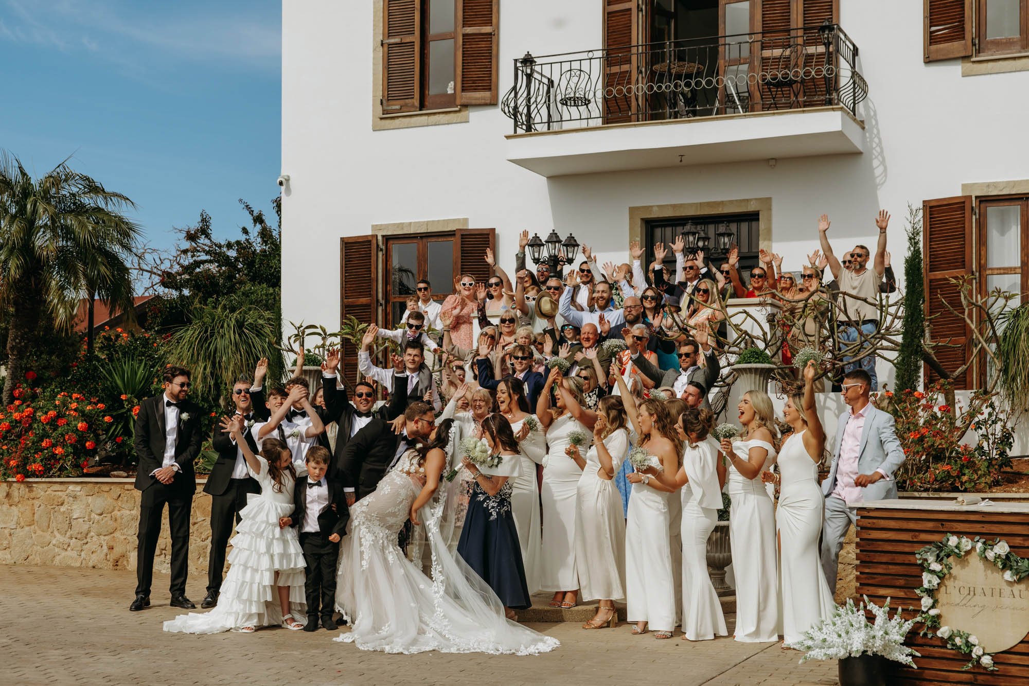 A large group of wedding guests and the bride and groom pose for a photo on the steps of a white building with wooden shutters. The bride is in a white wedding gown kissing a young boy, and the groom is in a black tuxedo. Many guests are dressed in f