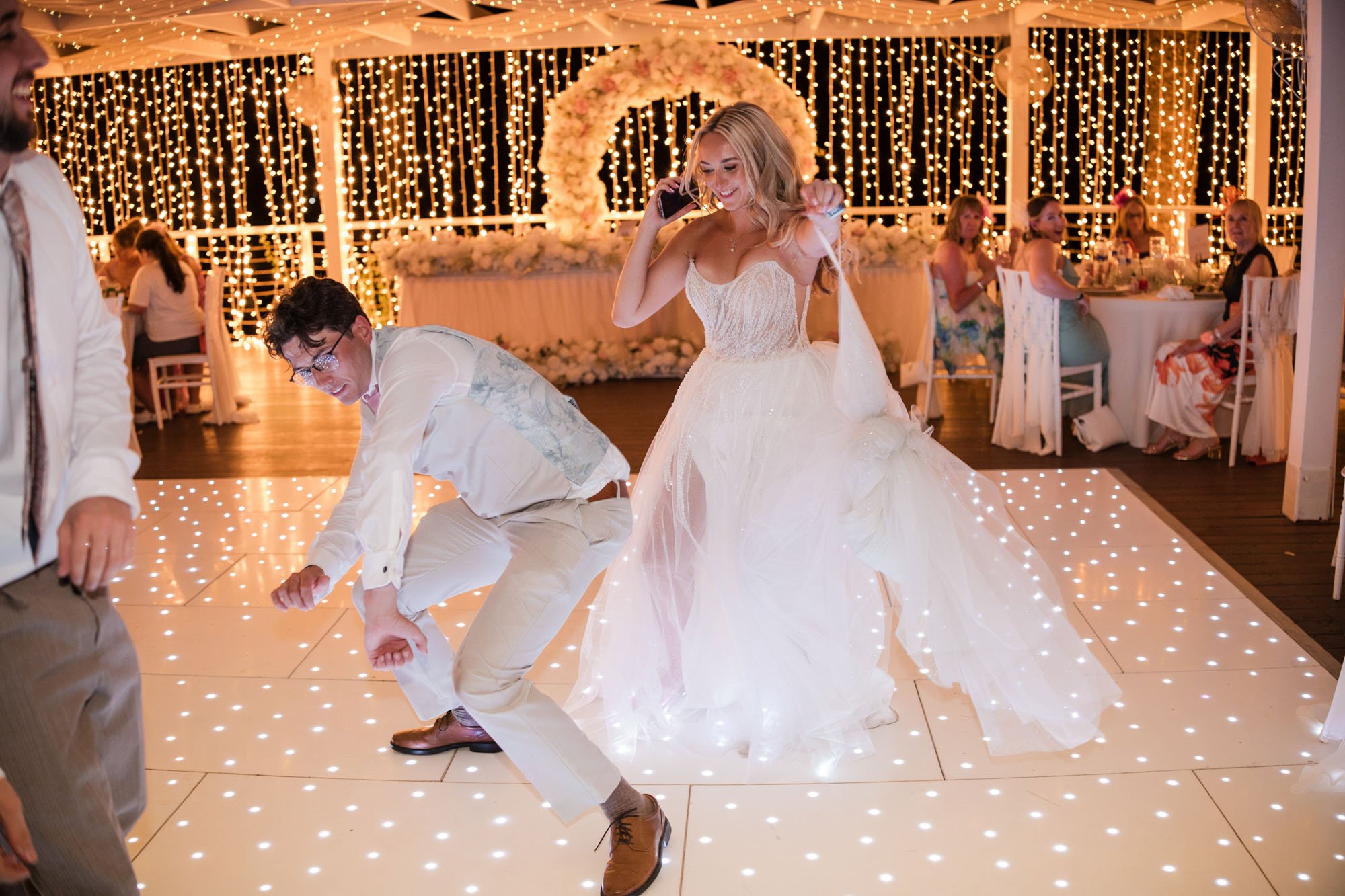 Wedding reception with a bride in a white gown dancing or celebrating on a lit dance floor, with guests seated at tables in the background decorated with string lights and floral arrangements.