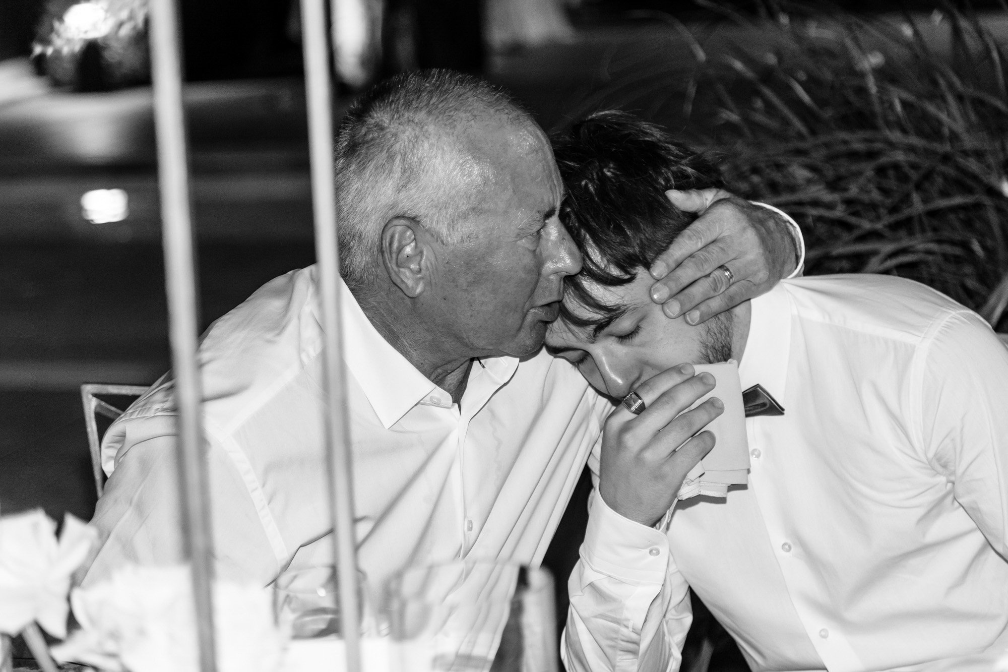 A man with short gray hair is comforting a young man with dark hair who is holding a drink, with their foreheads touching and one person's hand on the other's head, in a black and white photo.