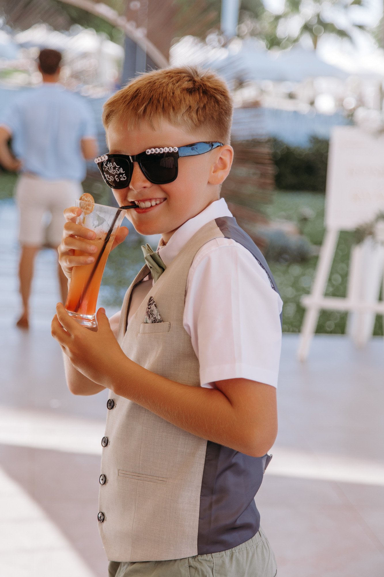 Smiling young boy in formal attire wearing large sunglasses and sipping a drink with a straw at an outdoor event.