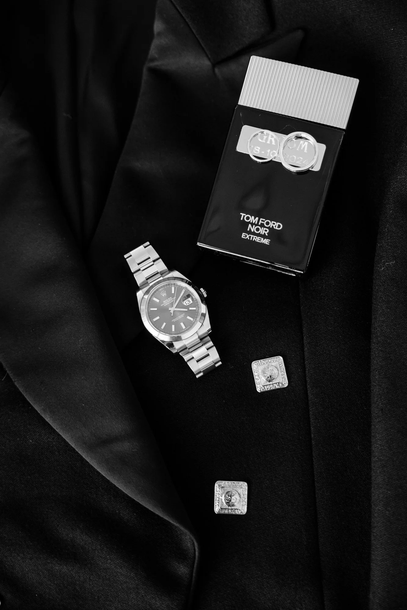 A wristwatch, a perfume box, a suit jacket, and a pair of cufflinks placed on a suit jacket.