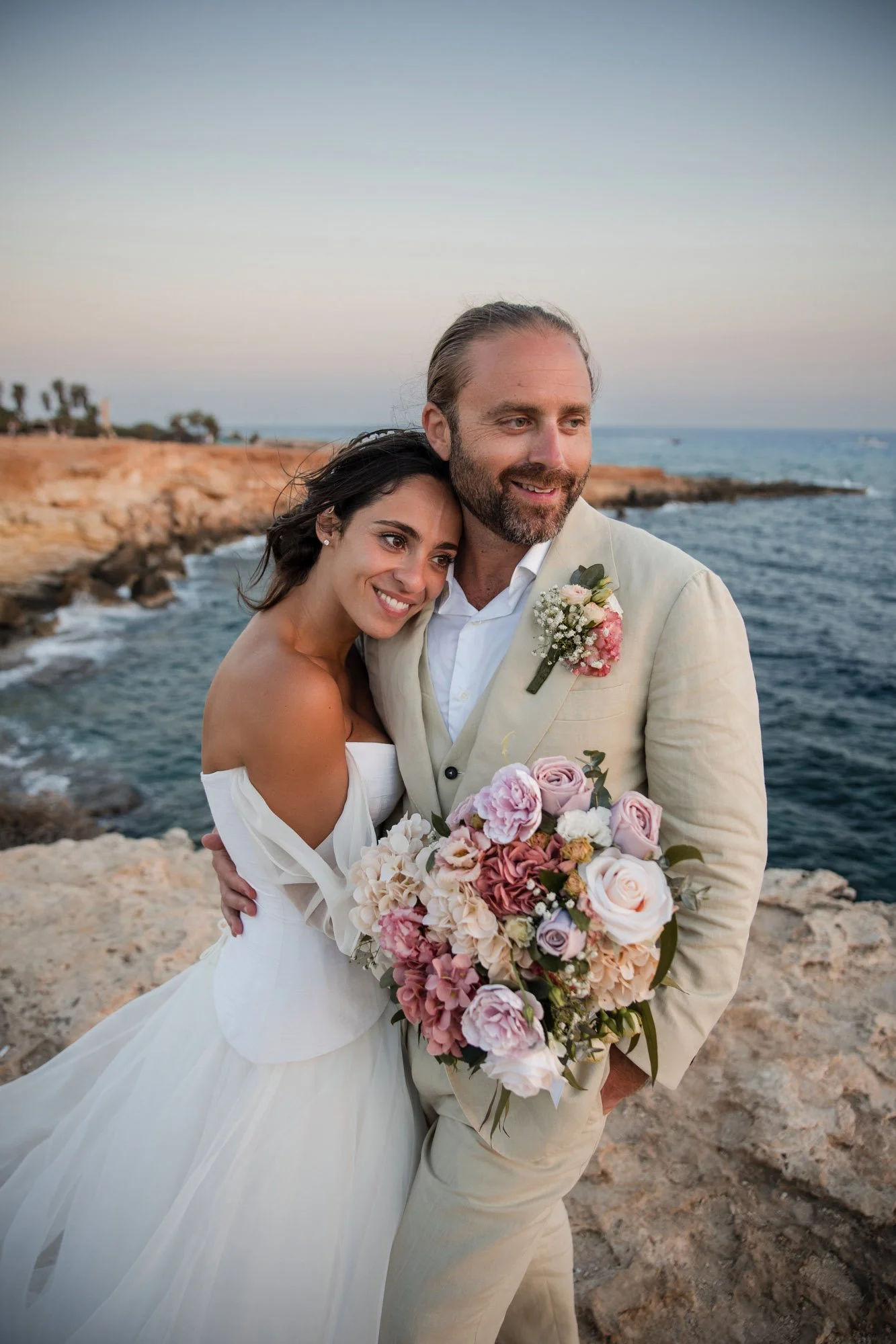 A bride and groom at the beach during sunset, smiling and holding a bouquet of flowers.