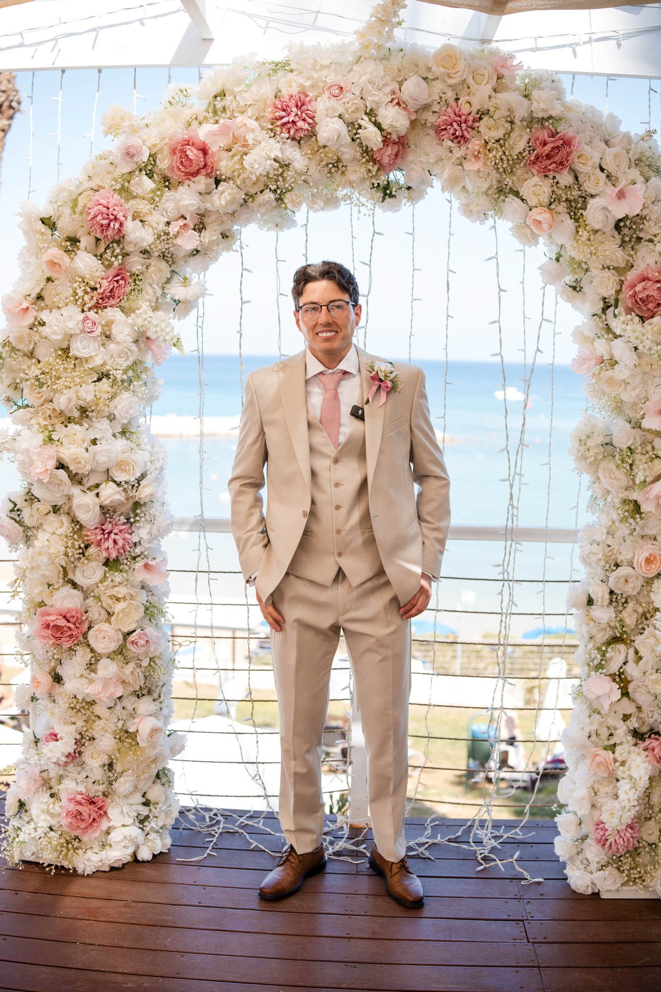 A man dressed in a cream-colored suit, pink tie, and glasses stands under a floral wedding arch with a beach and ocean in the background.