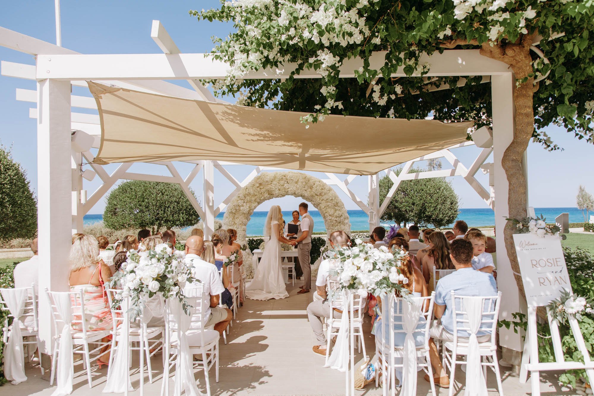A wedding ceremony taking place outdoors on a beachfront with white chairs, floral arrangements, and a floral arch, under a pergola with a beige shade sail, overlooking the ocean.