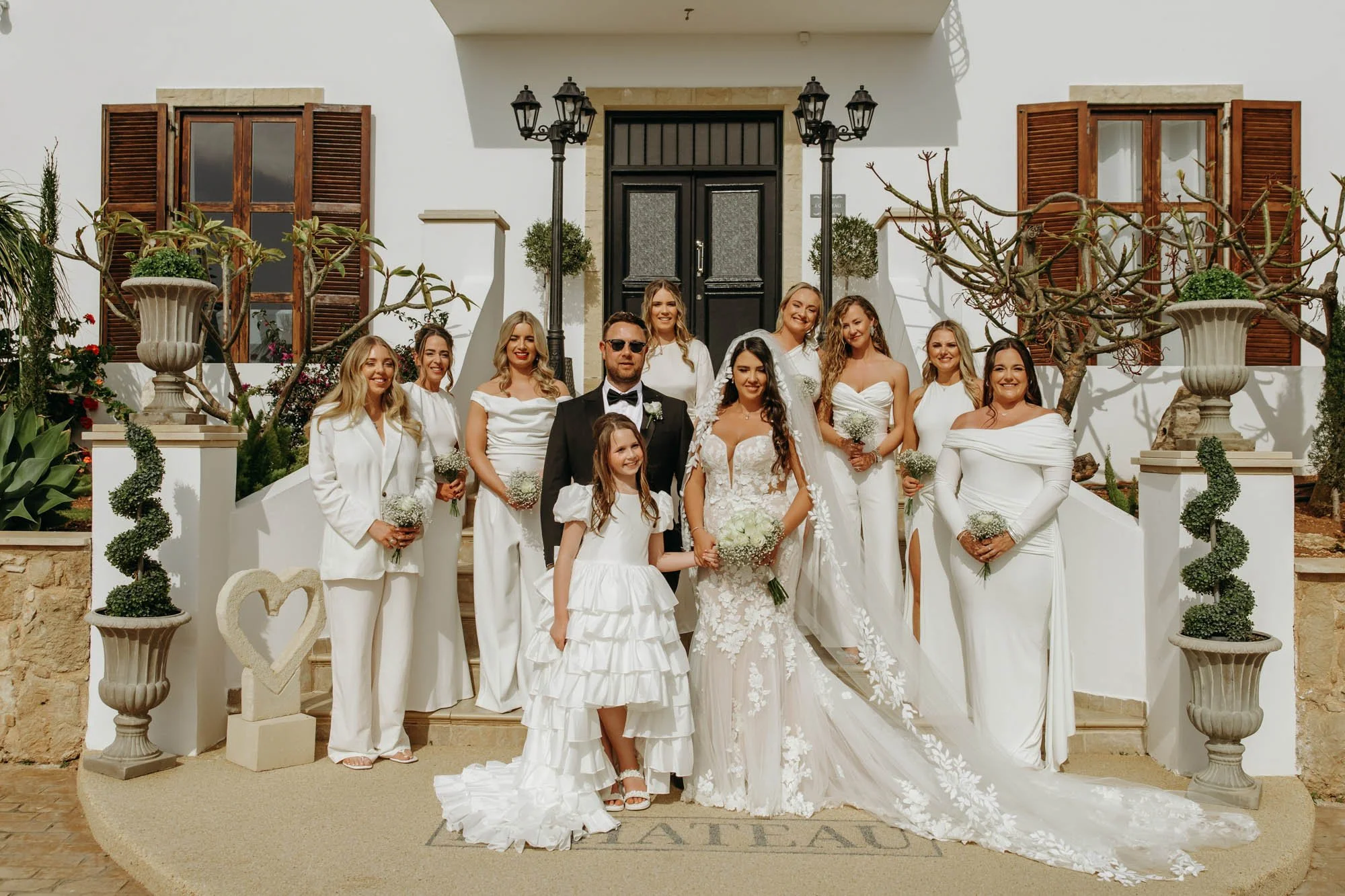 A wedding party in front of a white house with black door, featuring bride in lace wedding gown, groom in tuxedo, and ten women in white dresses, with a young girl in white dress holding the bride's hand.