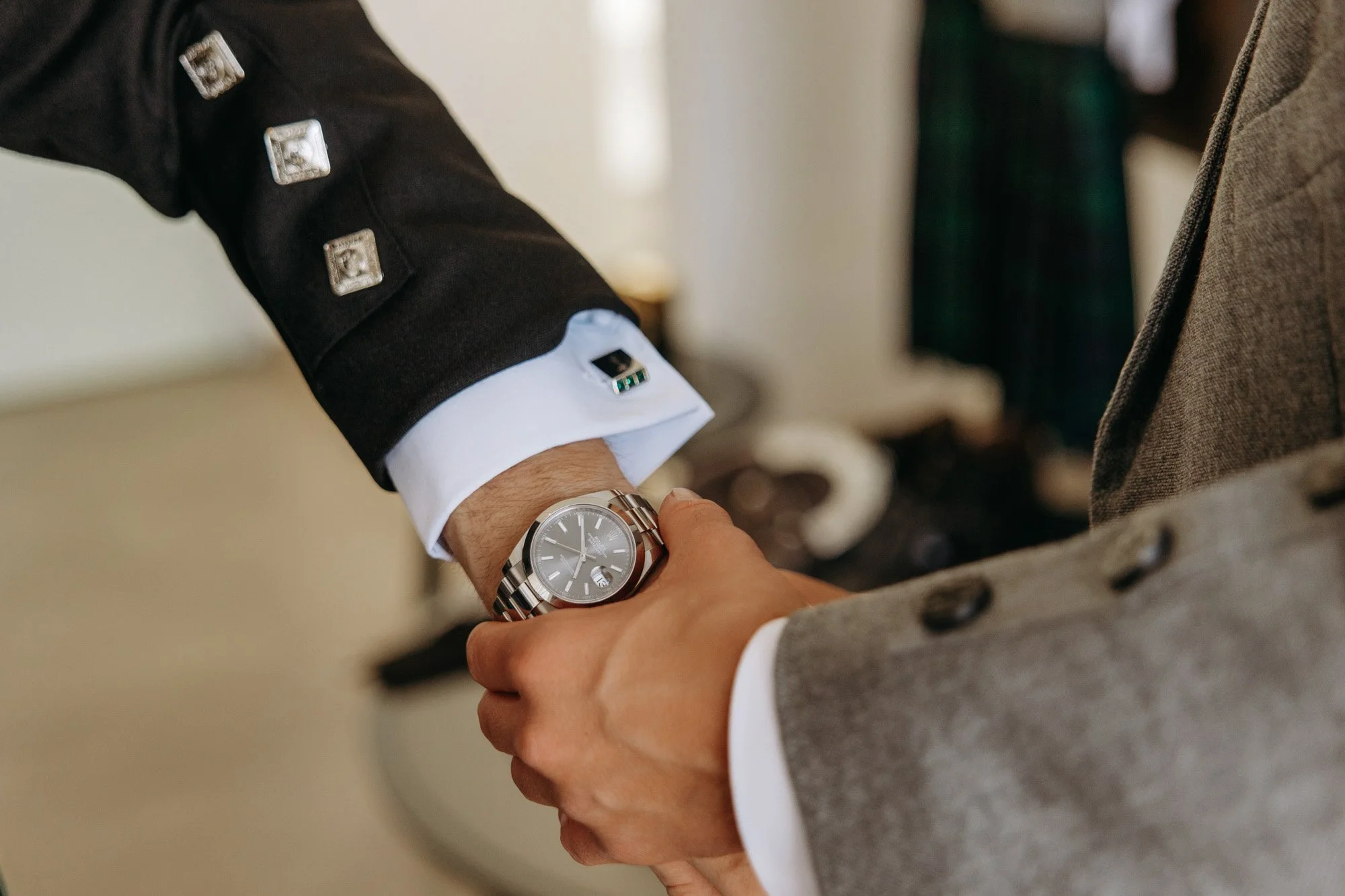 Two men shaking hands, with one wearing a watch and formal attire with a suit jacket and white shirt, and the other in a gray blazer.