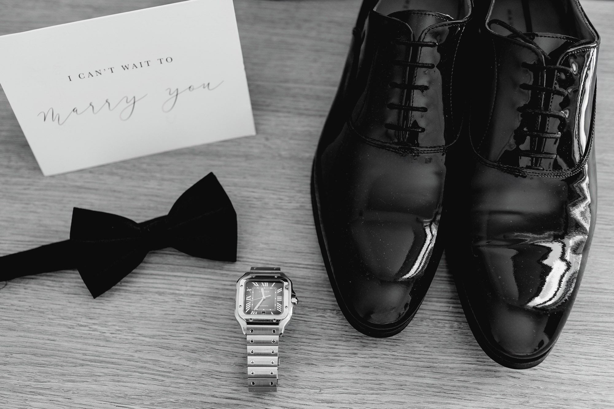 Black polished dress shoes, a wristwatch, a black bow tie, a white card with 'I CAN'T WAIT TO marry you' written on it, all arranged on a wooden surface.