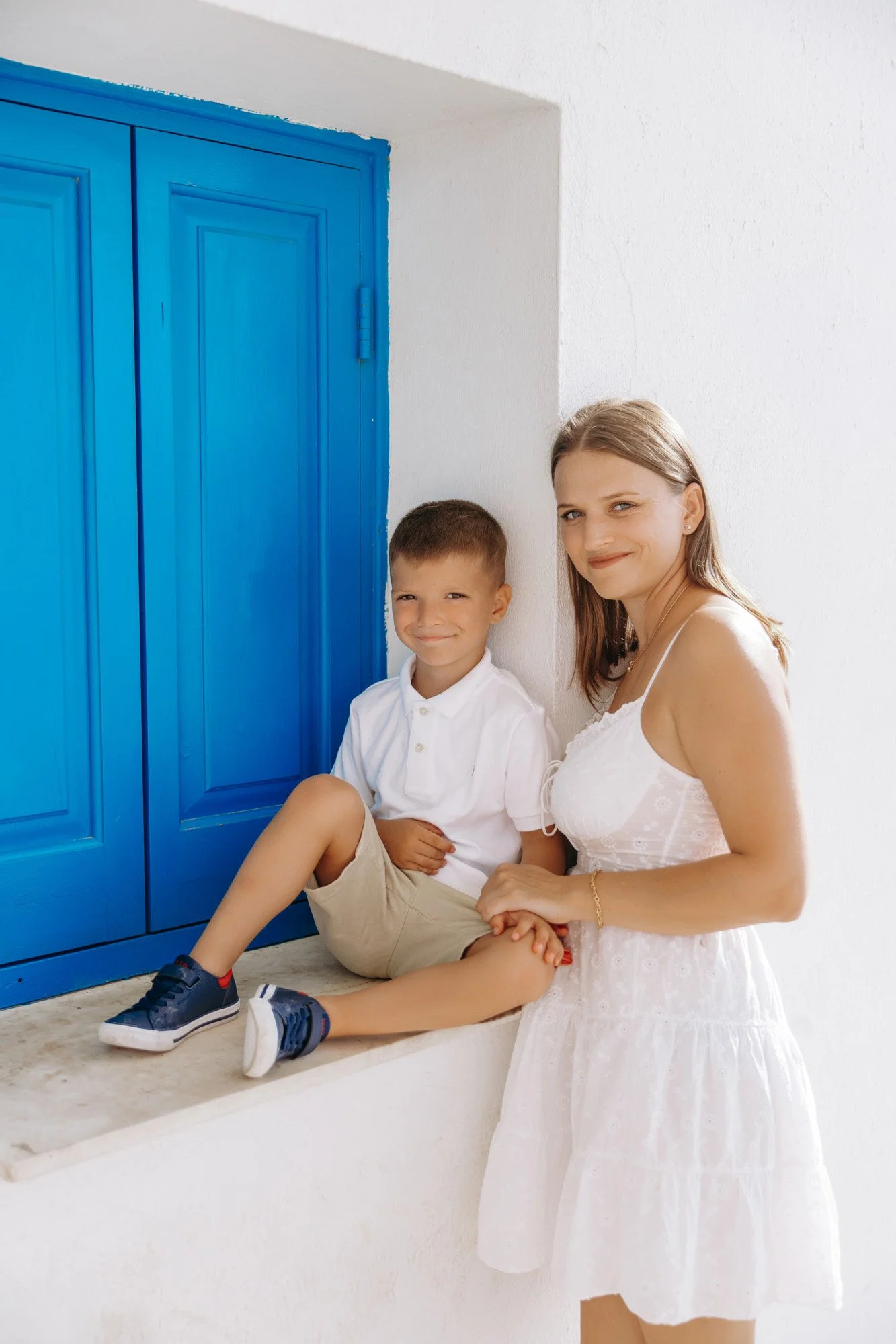 A woman in a white summer dress sitting on a ledge beside a young boy in a white shirt and khaki shorts, in front of a white wall and a blue door, both smiling.