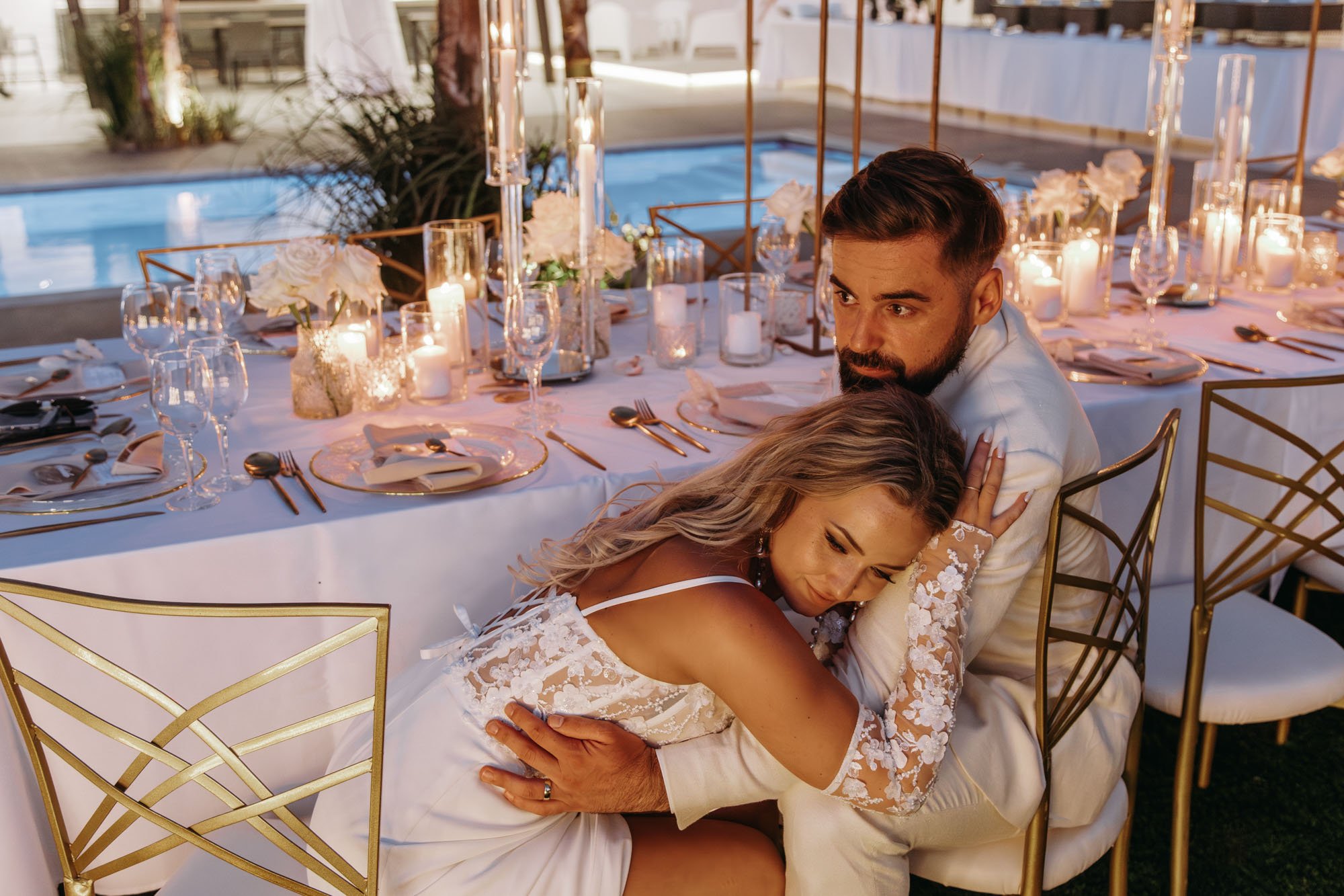 A couple embracing at a decorated outdoor dinner table during a wedding or special event, with candles and flowers.