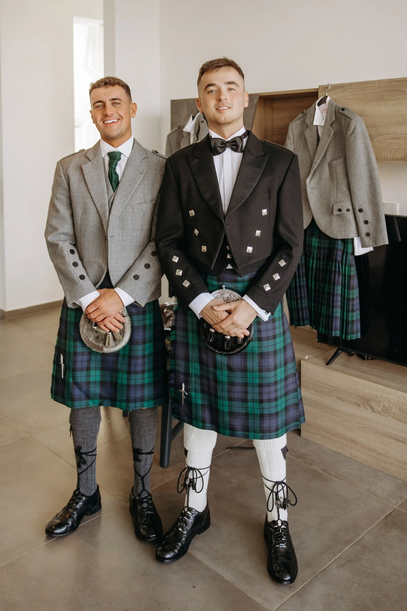 Two men dressed in traditional Scottish kilts and jackets, standing indoors. The man on the right wears a black jacket with silver embellishments and a black bow tie, while the man on the left wears a gray jacket with a green tie. Both are holding sp