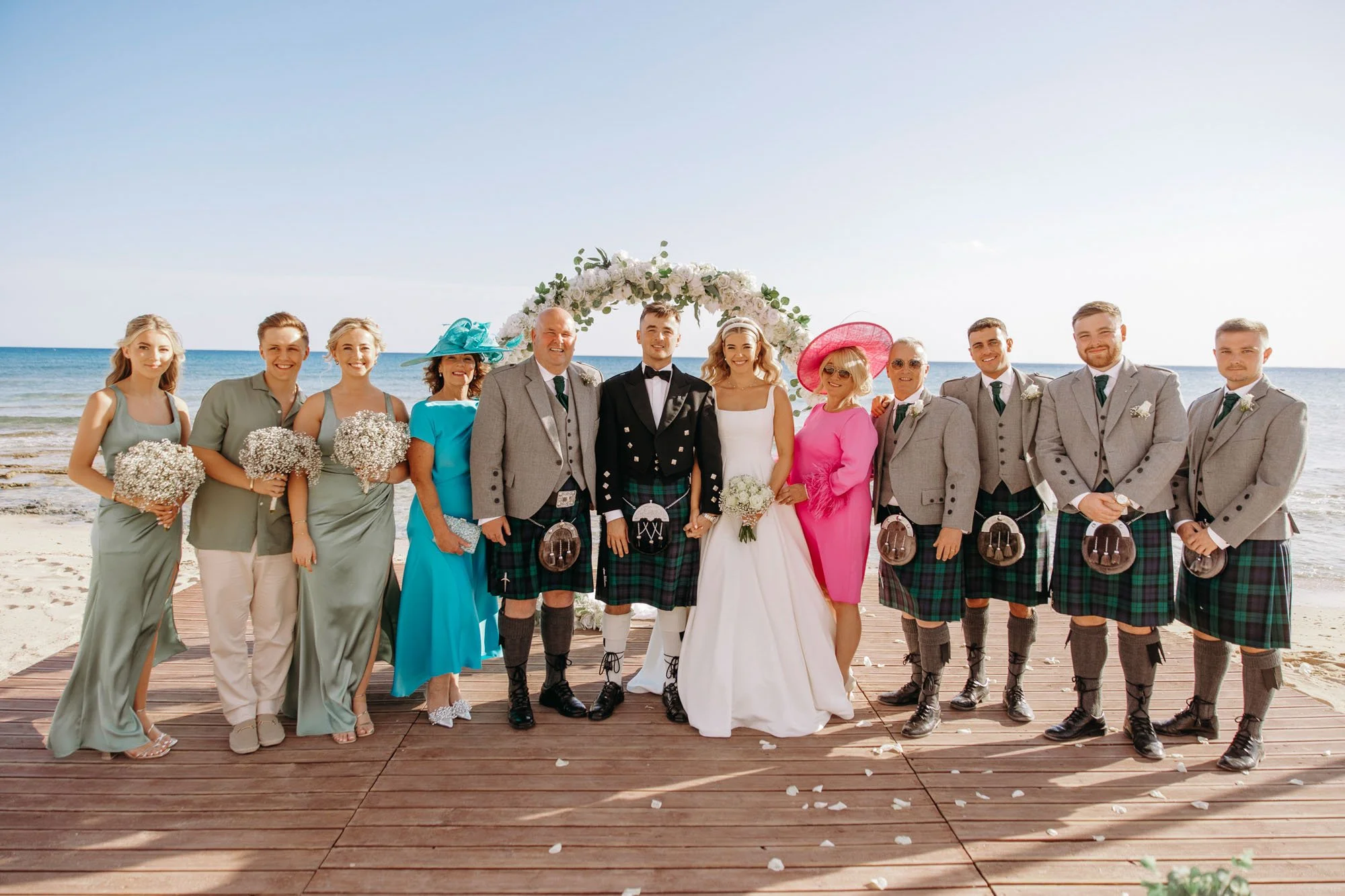 A group of people at a beach wedding, including the bride and groom, bridesmaids, groomsmen, and family members, standing on a wooden platform with the ocean in the background.