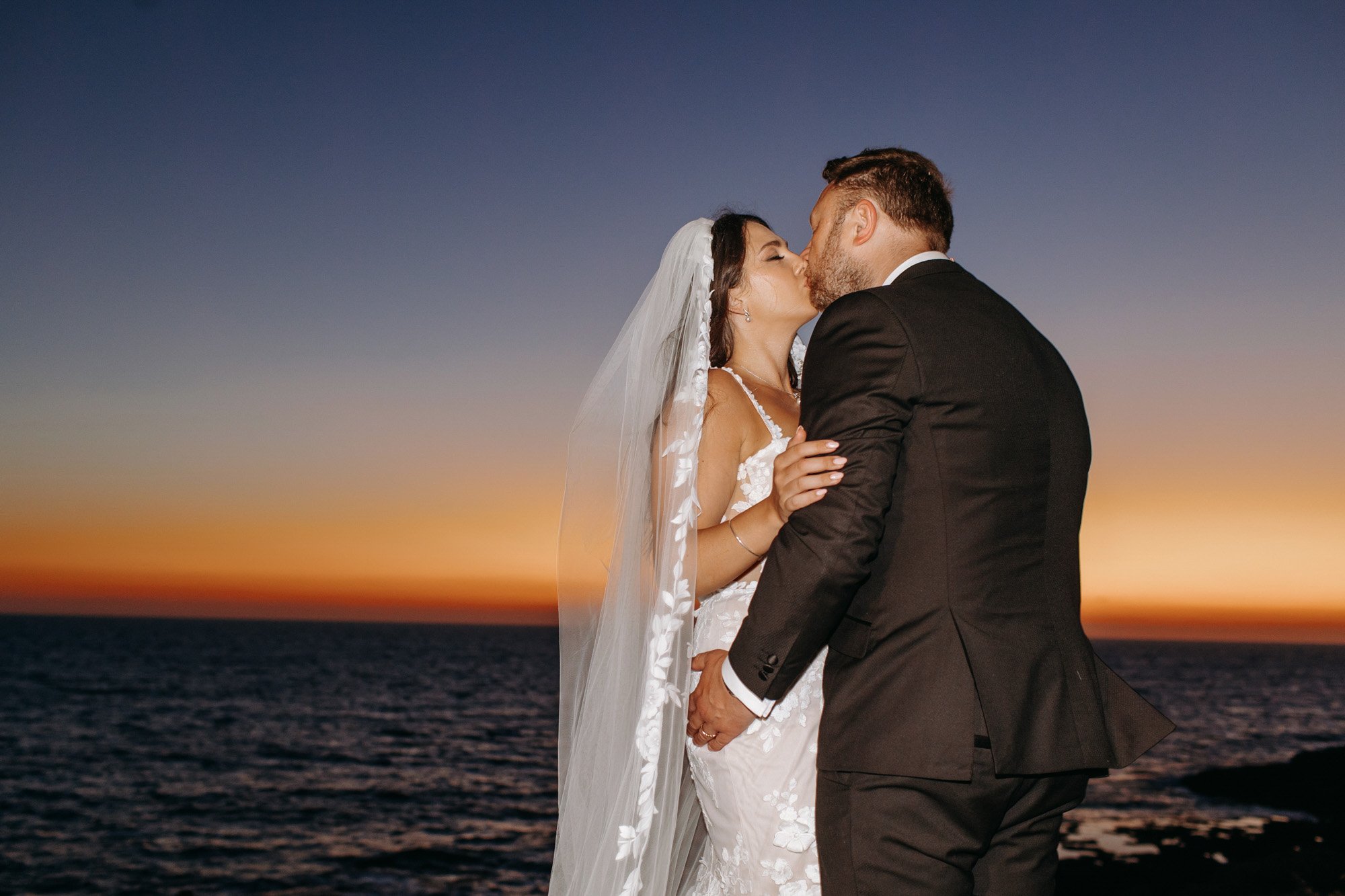 A bride and groom kissing during sunset by the ocean, with calm water and a colorful sky.