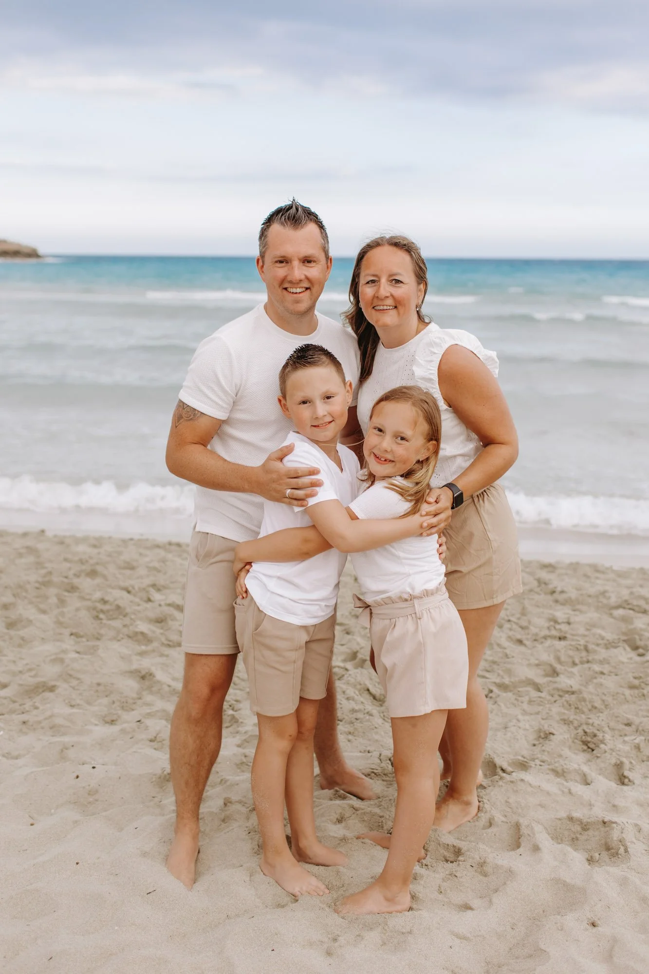 A family of four (a man, woman, and two children) standing on the beach with ocean waves and cloudy sky in the background. They are smiling and hugging.