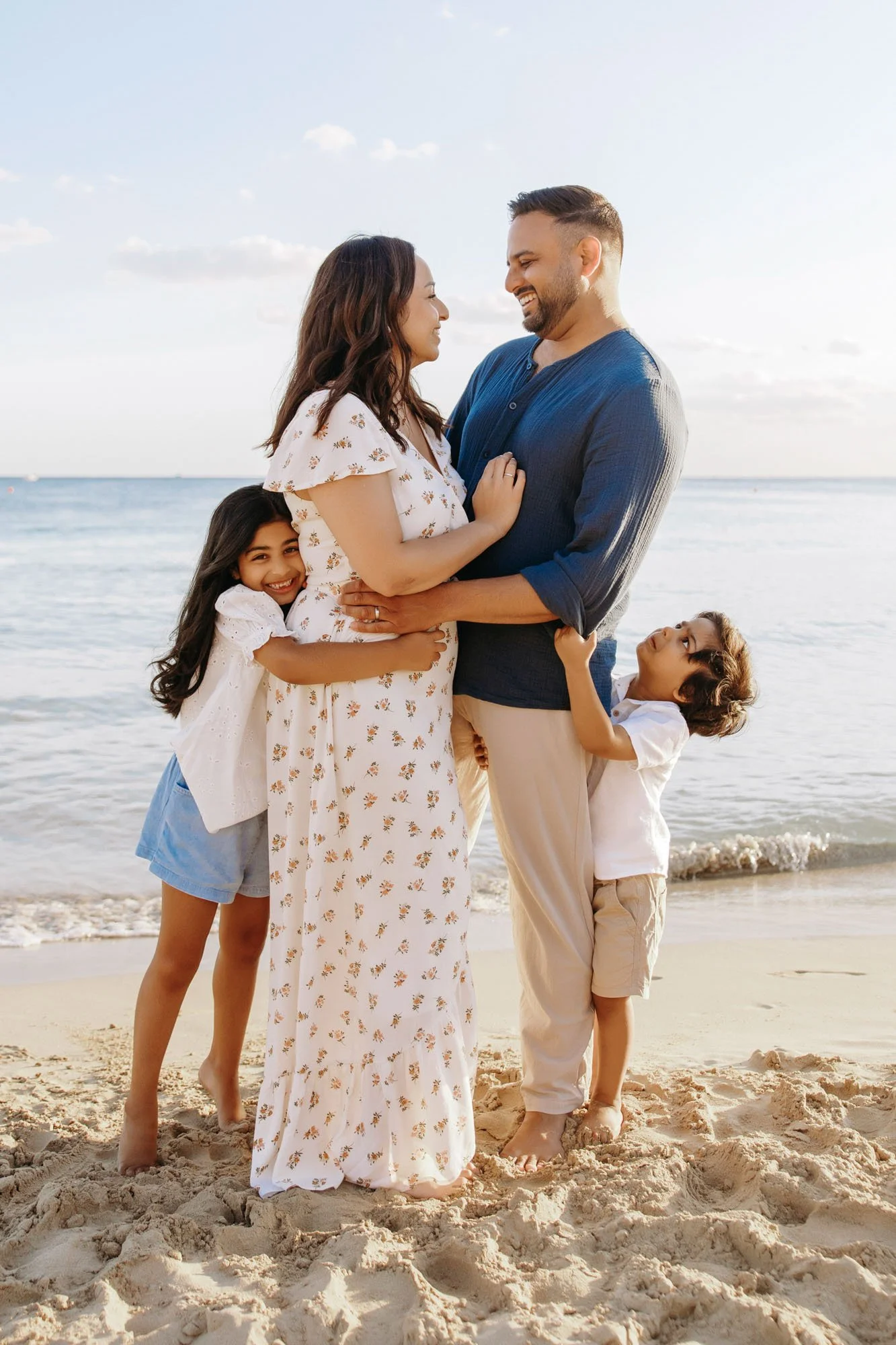 A family of four standing on a sandy beach near the ocean, smiling and looking at each other. The mother and father are in the center, with their children hugging them from the sides. The setting appears to be during sunset or late afternoon.