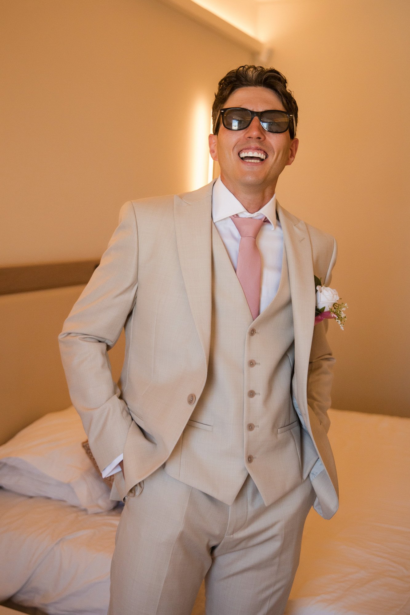 A man in a beige suit and pink tie smiling with sunglasses, standing near a bed in a warmly lit room.