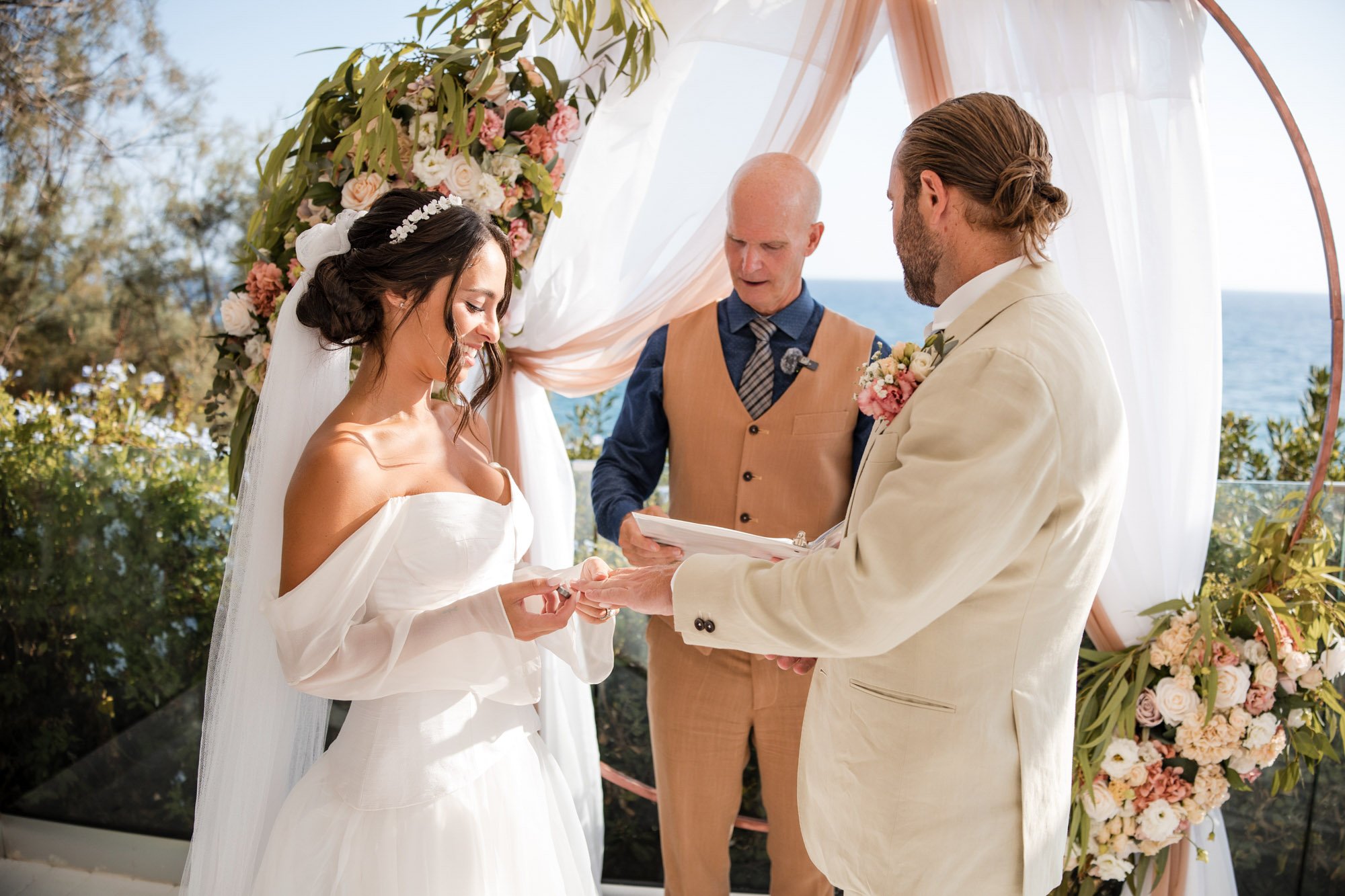 Bride and groom exchanging wedding rings under an outdoor arch decorated with pink and white flowers, with the ocean in the background.