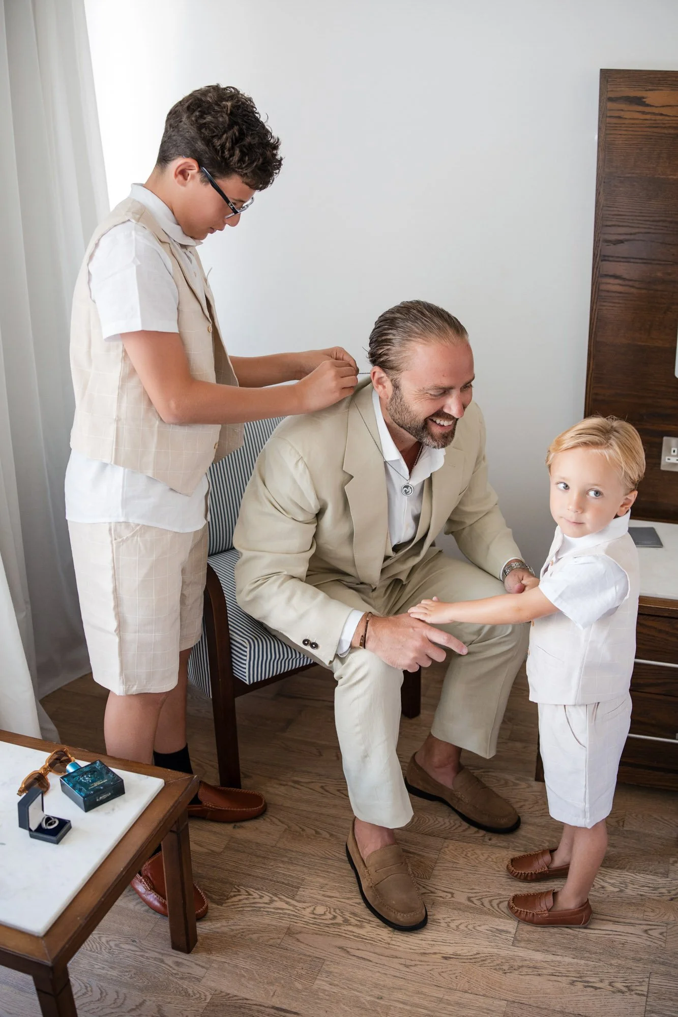 A father and two sons in matching white outfits, with the father sitting, one son fixing his hair, and the other son holding his hand.