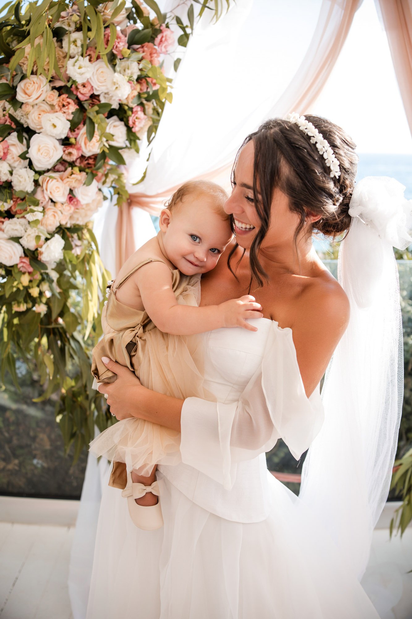 A bride holding a young girl, possibly a flower girl, both smiling happily. The bride is wearing a white wedding dress with an off-the-shoulder style and a floral headband. The background features floral decorations and sheer curtains, with a view of