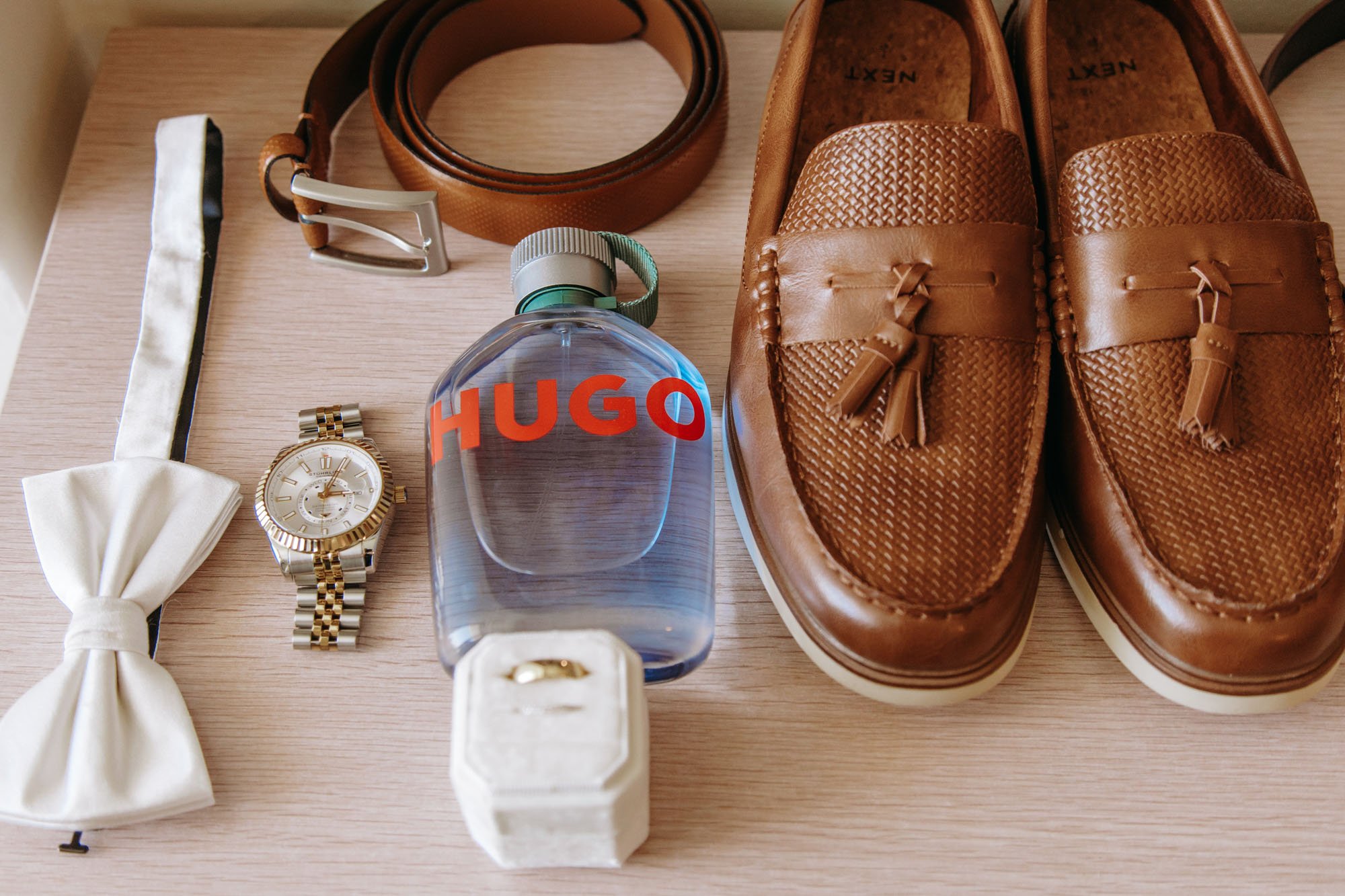 Collection of men's accessories and footwear on a wooden surface, including brown loafers, a silver and gold wristwatch, a white bow tie, a leather belt, a bottle of Hugo cologne, and a ring in a jewelry box.