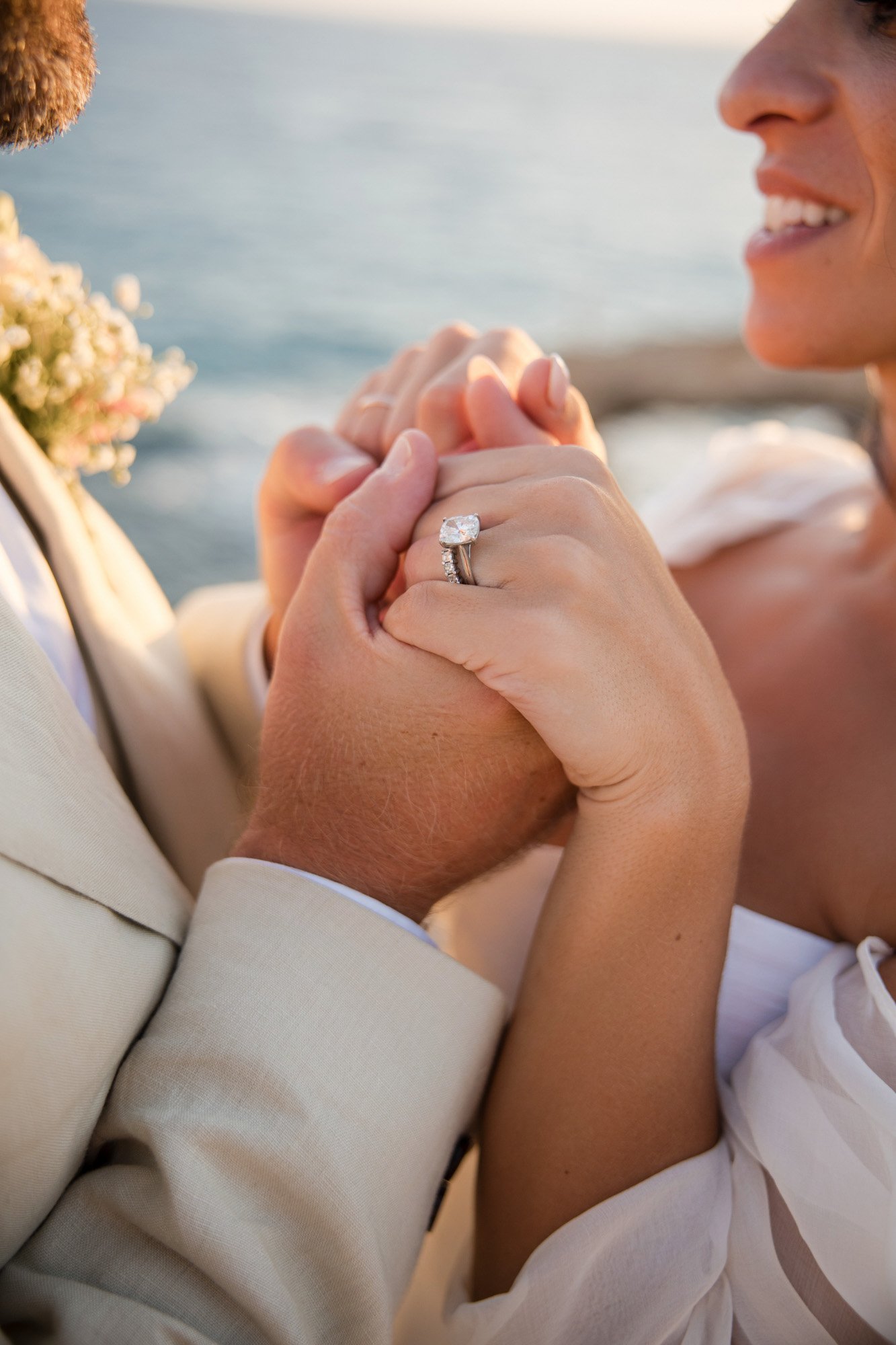 Close-up of a couple holding hands, showing an engagement ring, with a body of water in the background.