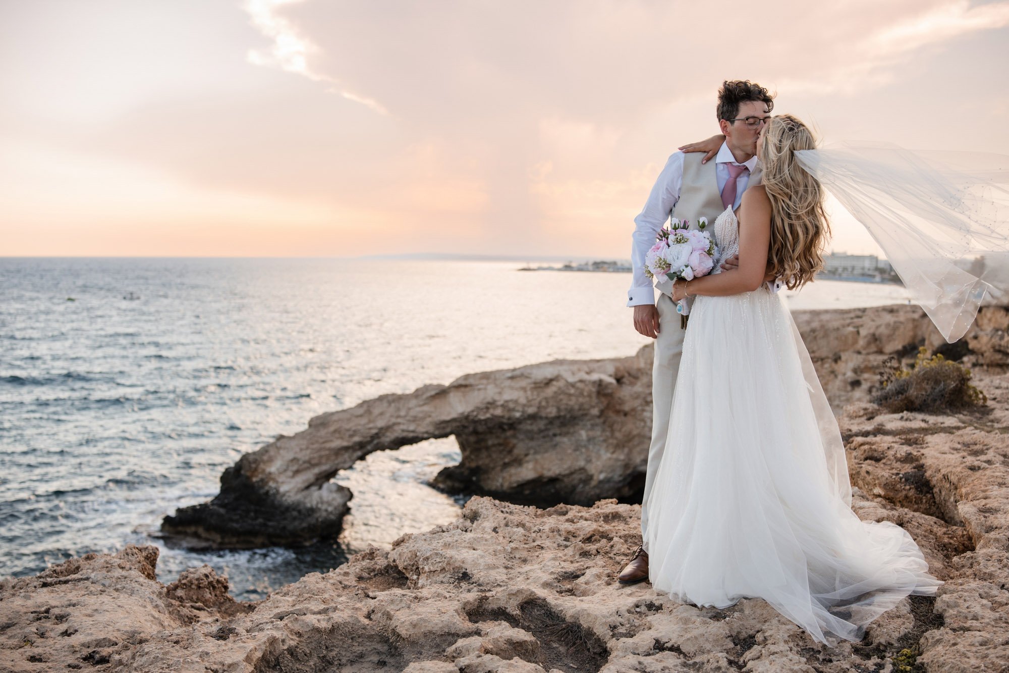 A bride and groom kissing on rocky cliffs by the ocean at sunset, with the bride holding a bouquet of flowers and wearing a flowing white wedding dress and veil.