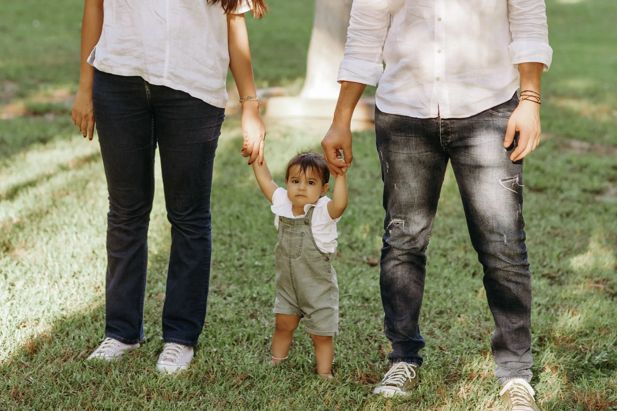 A toddler holding hands with two adults, one on each side, in a grassy park.