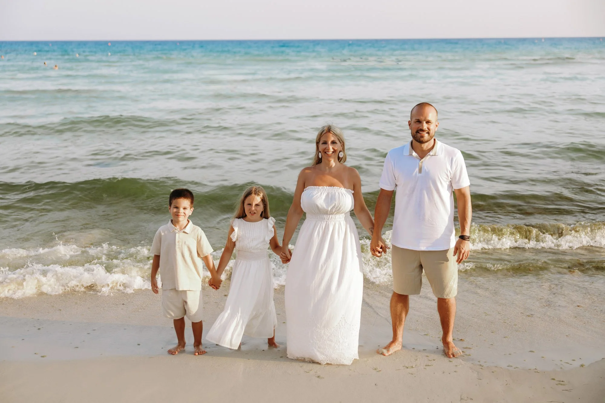 A family of five holding hands at the beach, with ocean waves in the background. The mother is wearing a white dress, the father a white polo shirt and beige shorts, and three children in light-colored clothing.