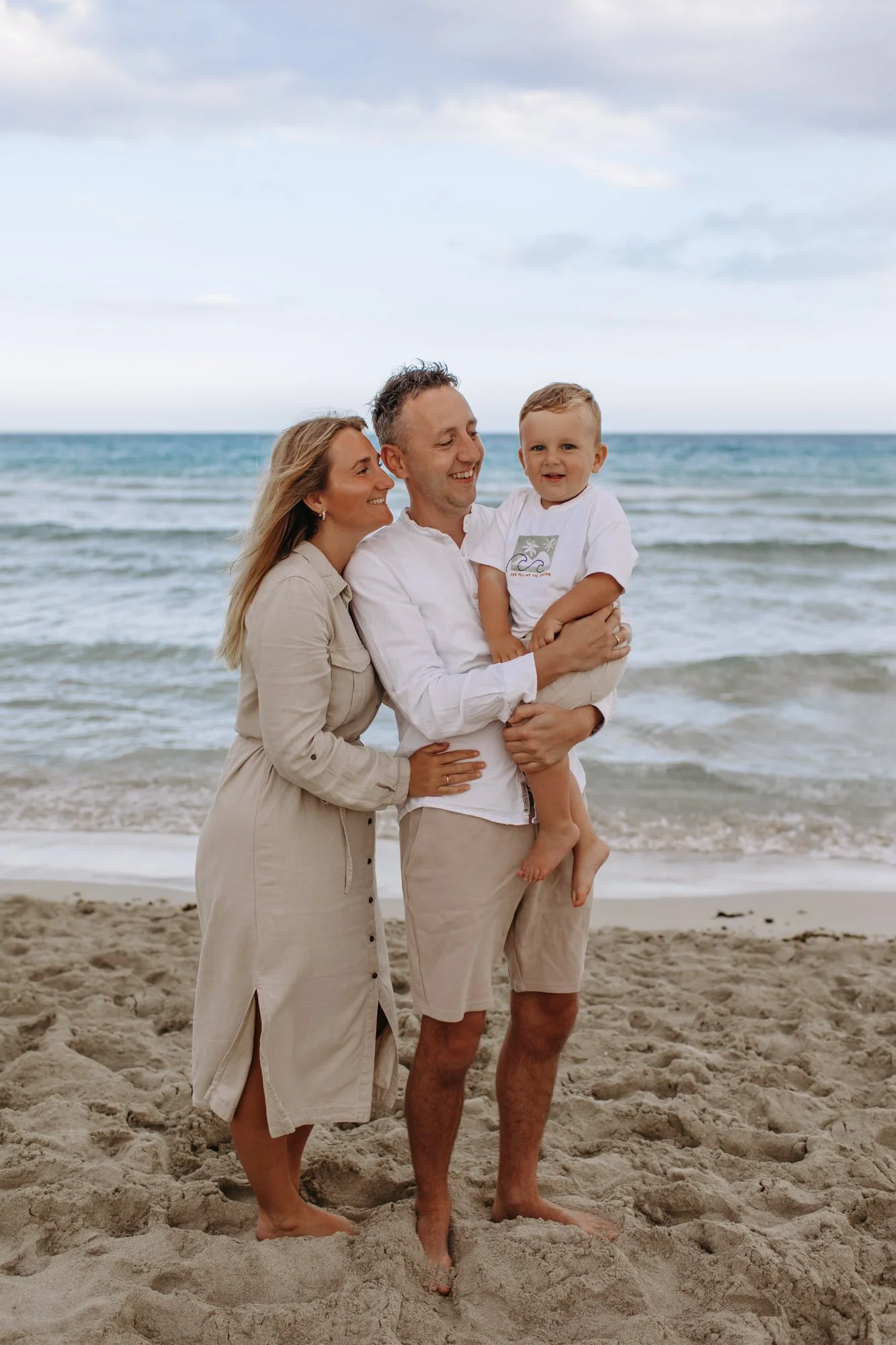A happy family of three on the beach, with the man holding a young boy, and the woman standing beside them, all smiling. The family is dressed in casual, light-colored clothes, and the ocean and cloudy sky are in the background.