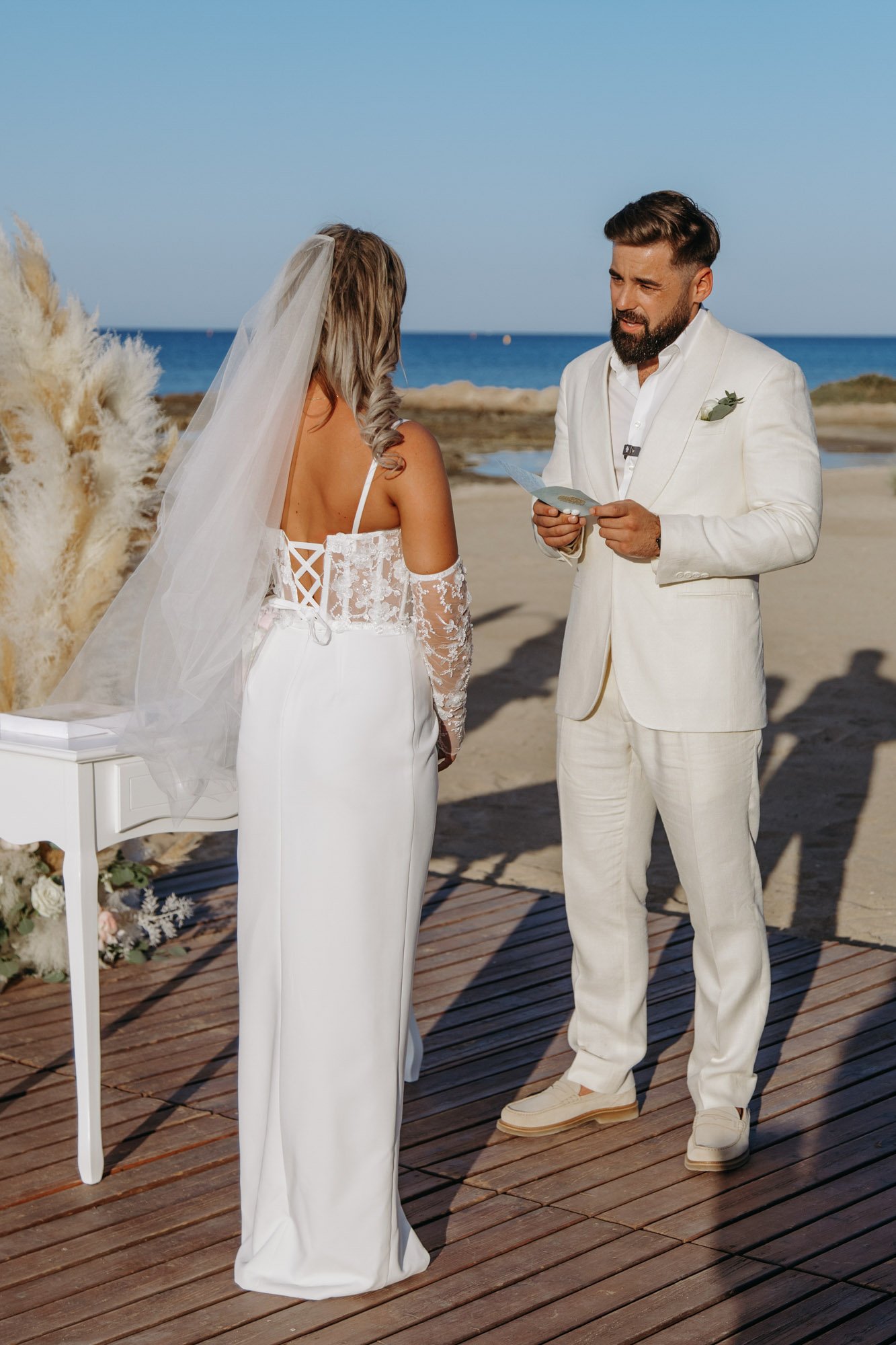 A couple getting married on the beach; the bride in a white gown with sheer sleeves and veil, the groom in a white suit, exchanging vows by the ocean with a wedding officiant reading from a paper.