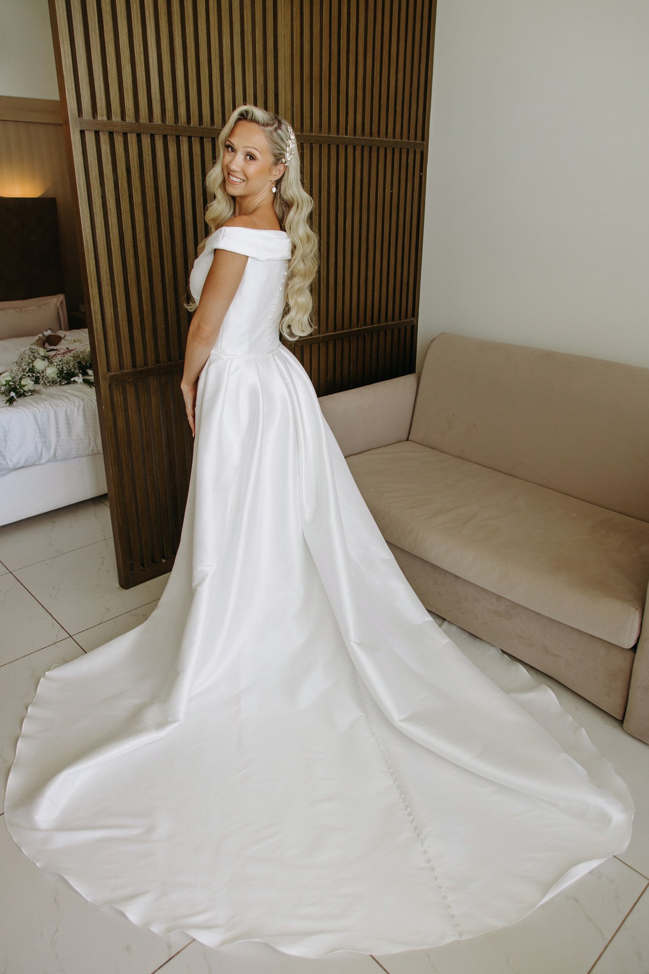 A bride in a white wedding dress standing indoors, smiling at the camera, with a bedroom with floral decor visible in the background.