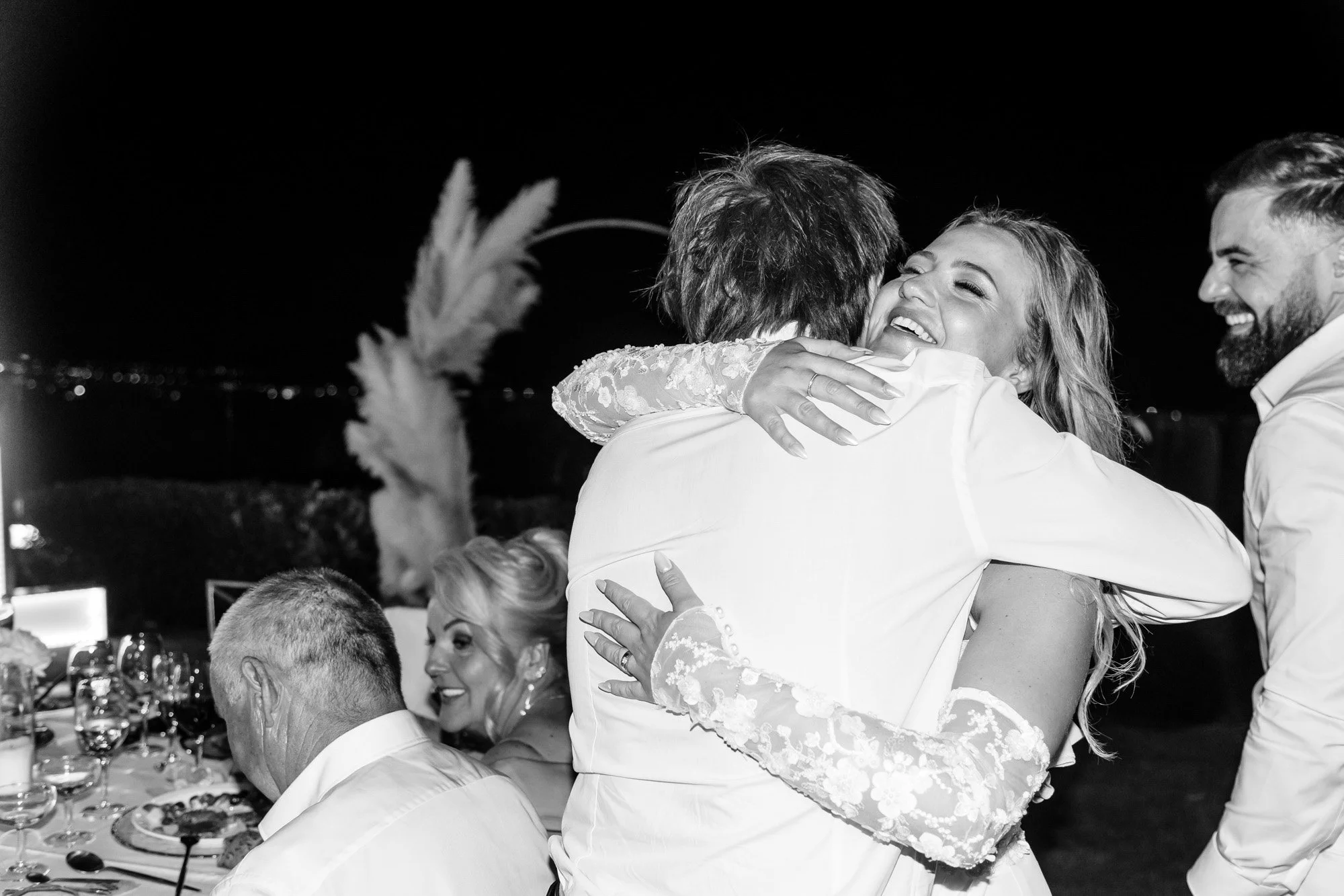 A woman in a wedding dress hugging a man in a white shirt at a wedding reception, with other guests smiling and enjoying the event.