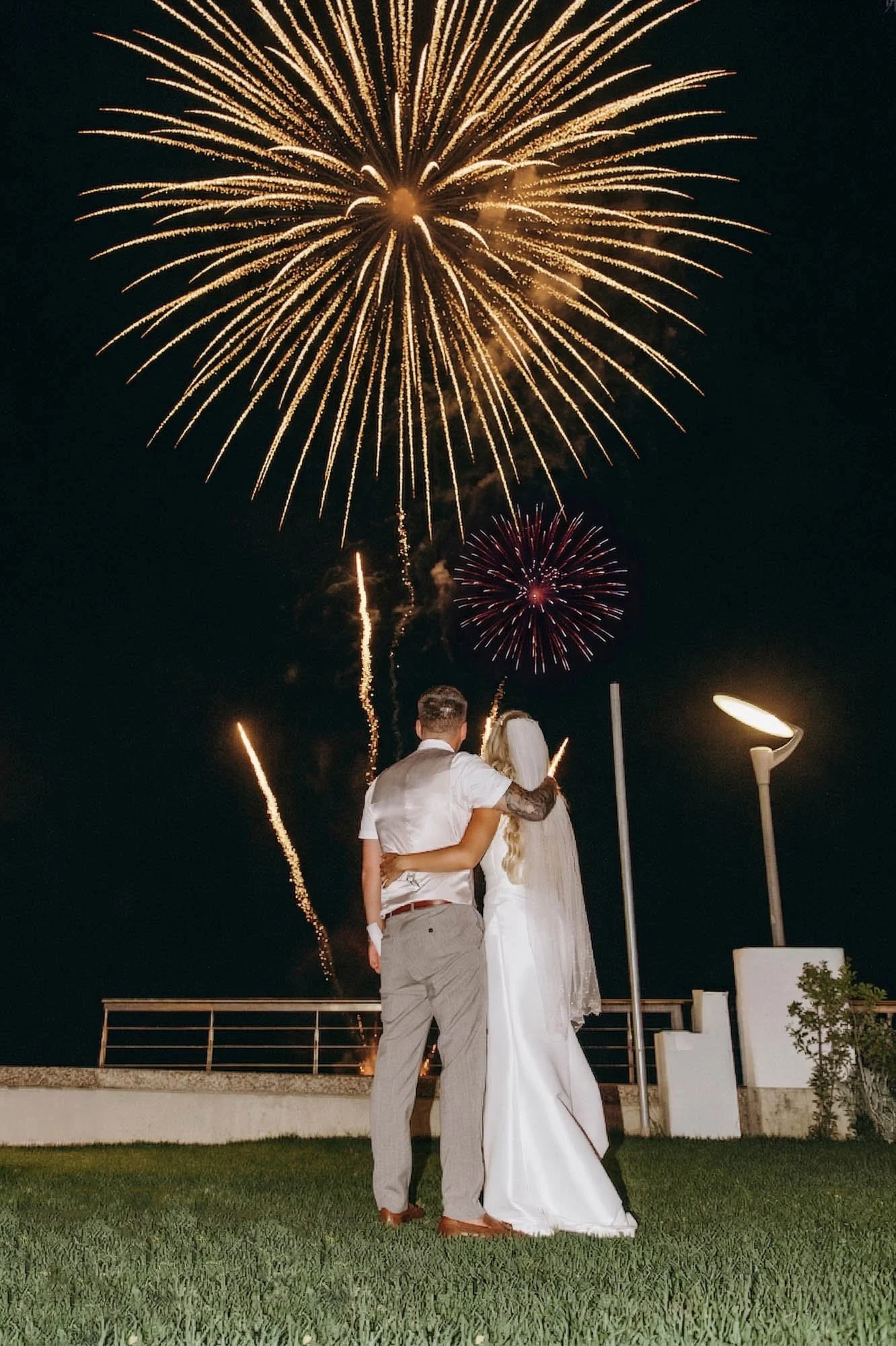 A couple dressed in wedding attire watching fireworks display at night.