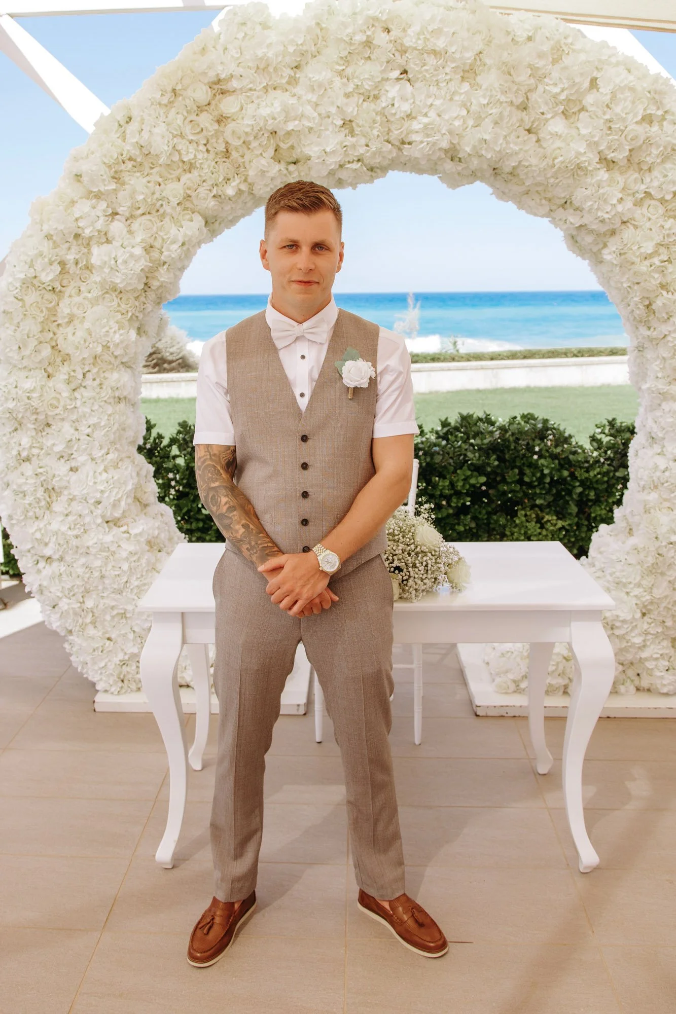 A man in wedding attire stands in front of a large floral heart with a beach and ocean view in the background, outdoors during daytime.