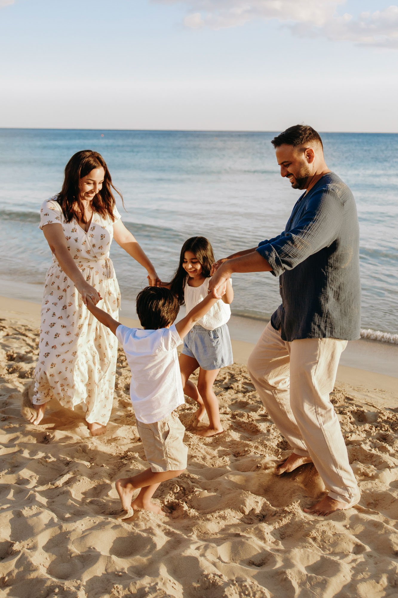 A family of four playing and holding hands on a sandy beach near the ocean, with clear blue water and a partly cloudy sky in the background.