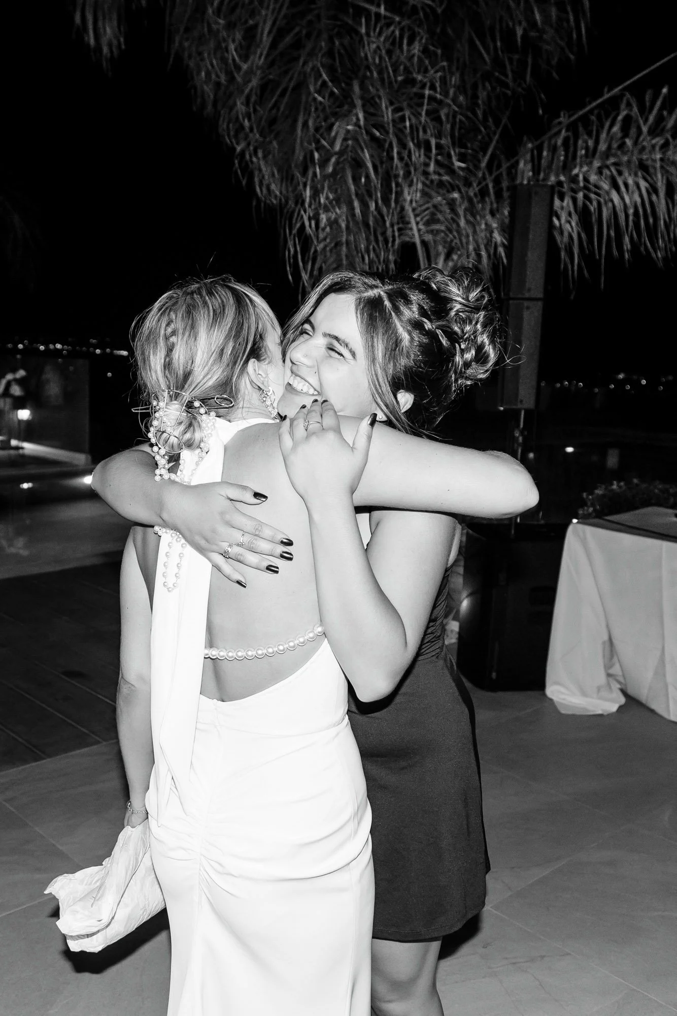Two women hugging and smiling at night, one in a white dress and the other in a dark dress, with palm trees and outdoor setting in the background.