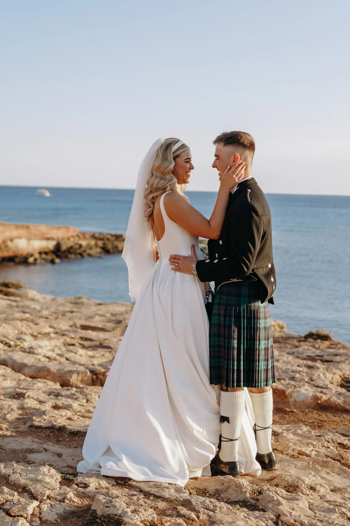 A bride and groom embrace on a rocky shoreline with the ocean and a boat in the background, during sunset.