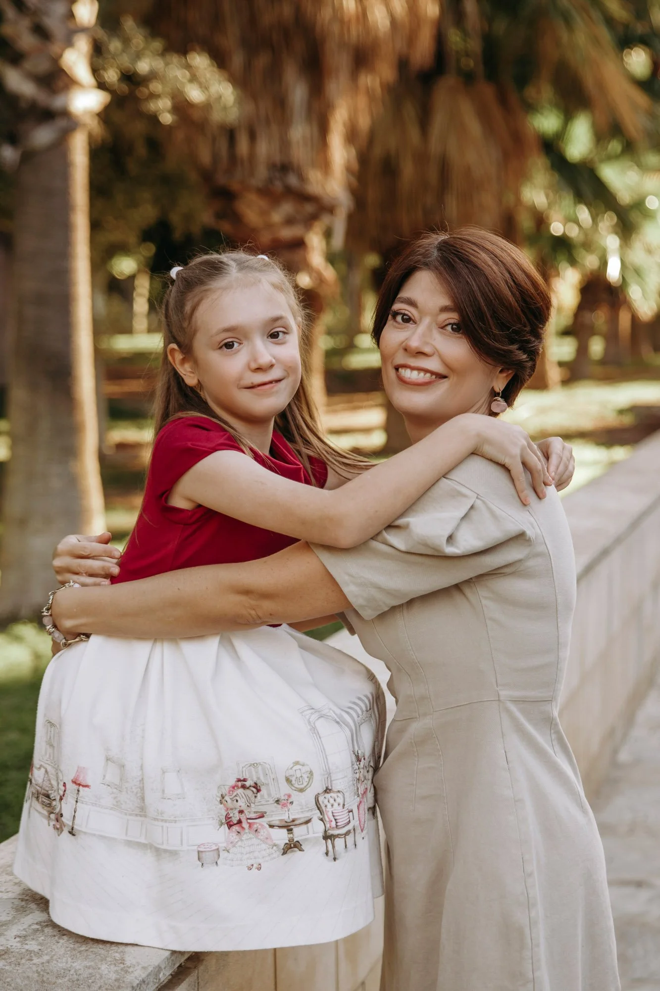 A woman with short brown hair and a girl with long light brown hair are embracing outdoors, with trees in the background.