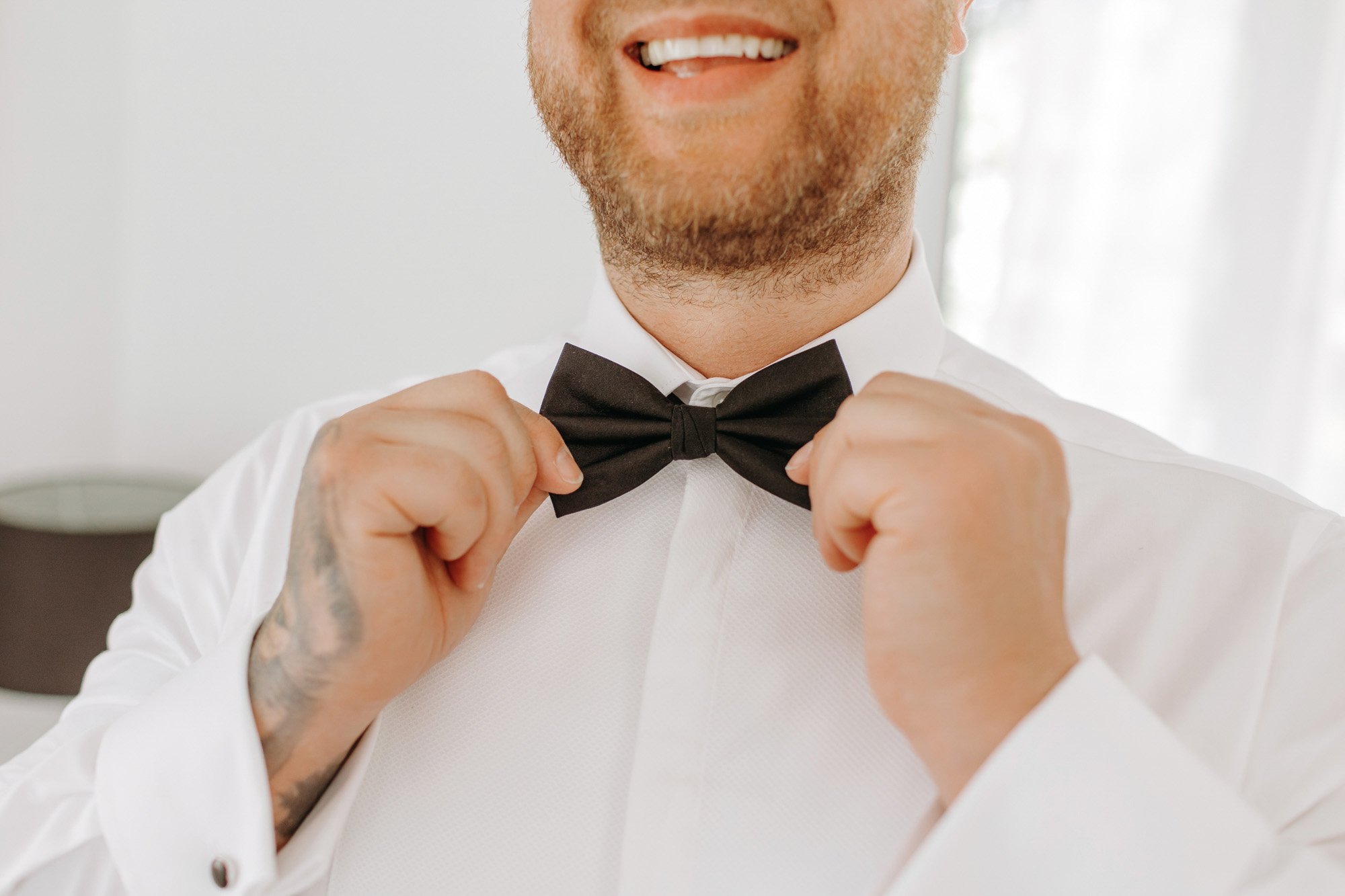 A man adjusting his black bow tie, smiling, wearing a white dress shirt, with a beard and tattooed hand, indoors with white curtains in the background.