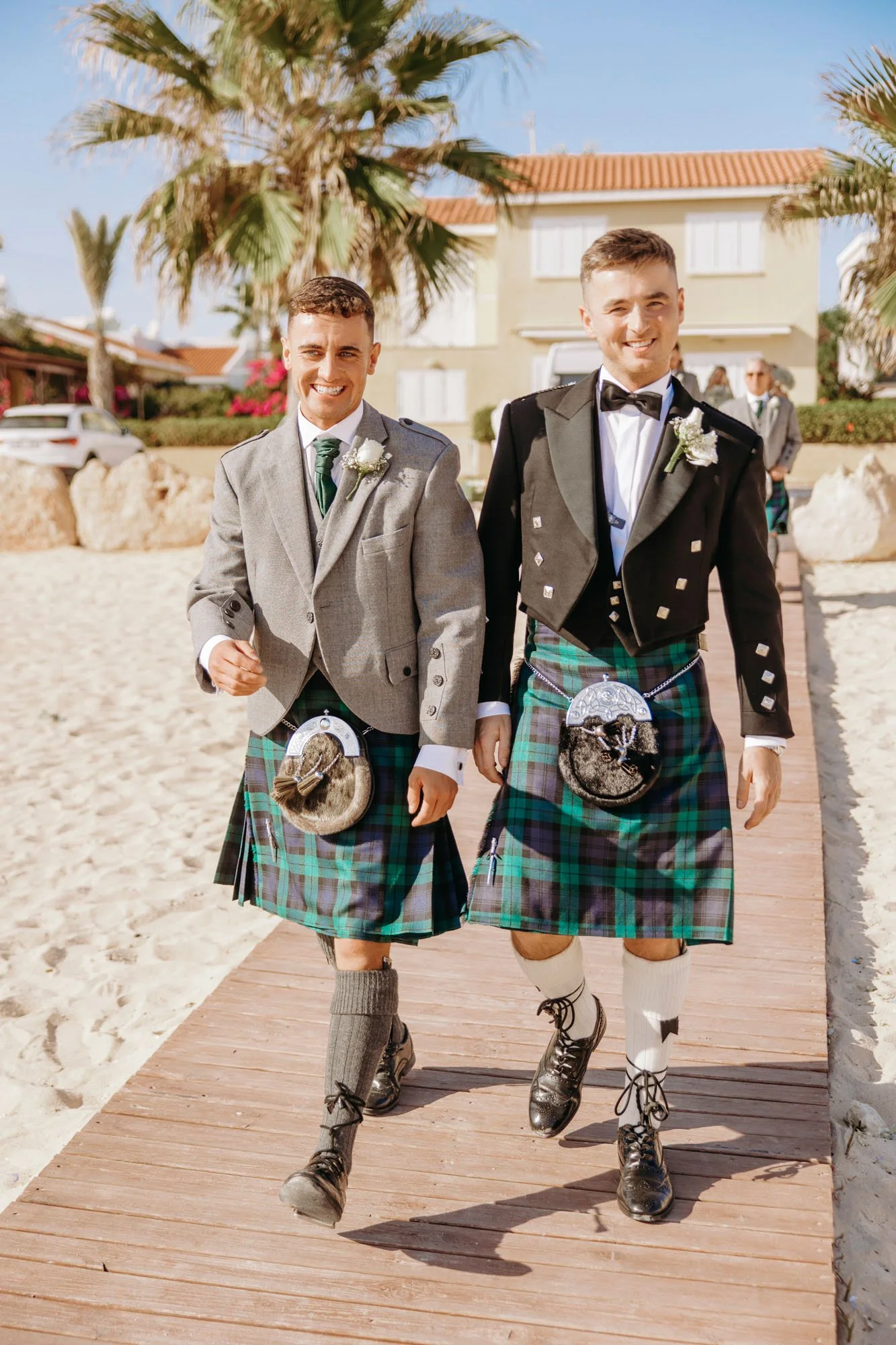 Two men wearing traditional Scottish kilts and jackets, walking on a wooden pathway at a beachside location, smiling and dressed for a wedding or special event.