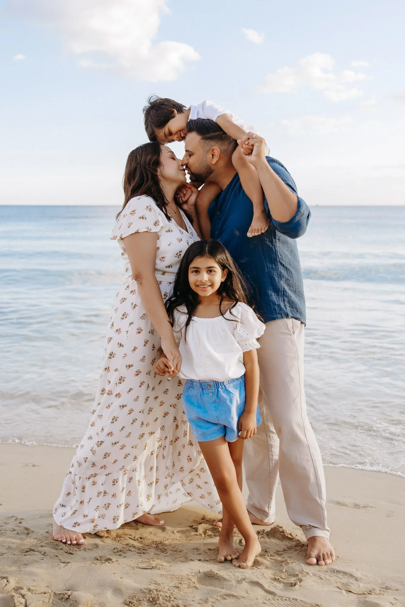 A family of four enjoying time together on the beach, with a man and woman kissing, a young girl smiling, and a young boy playfully lying on a man's shoulders, all barefoot on the sand with the ocean and sky in the background.