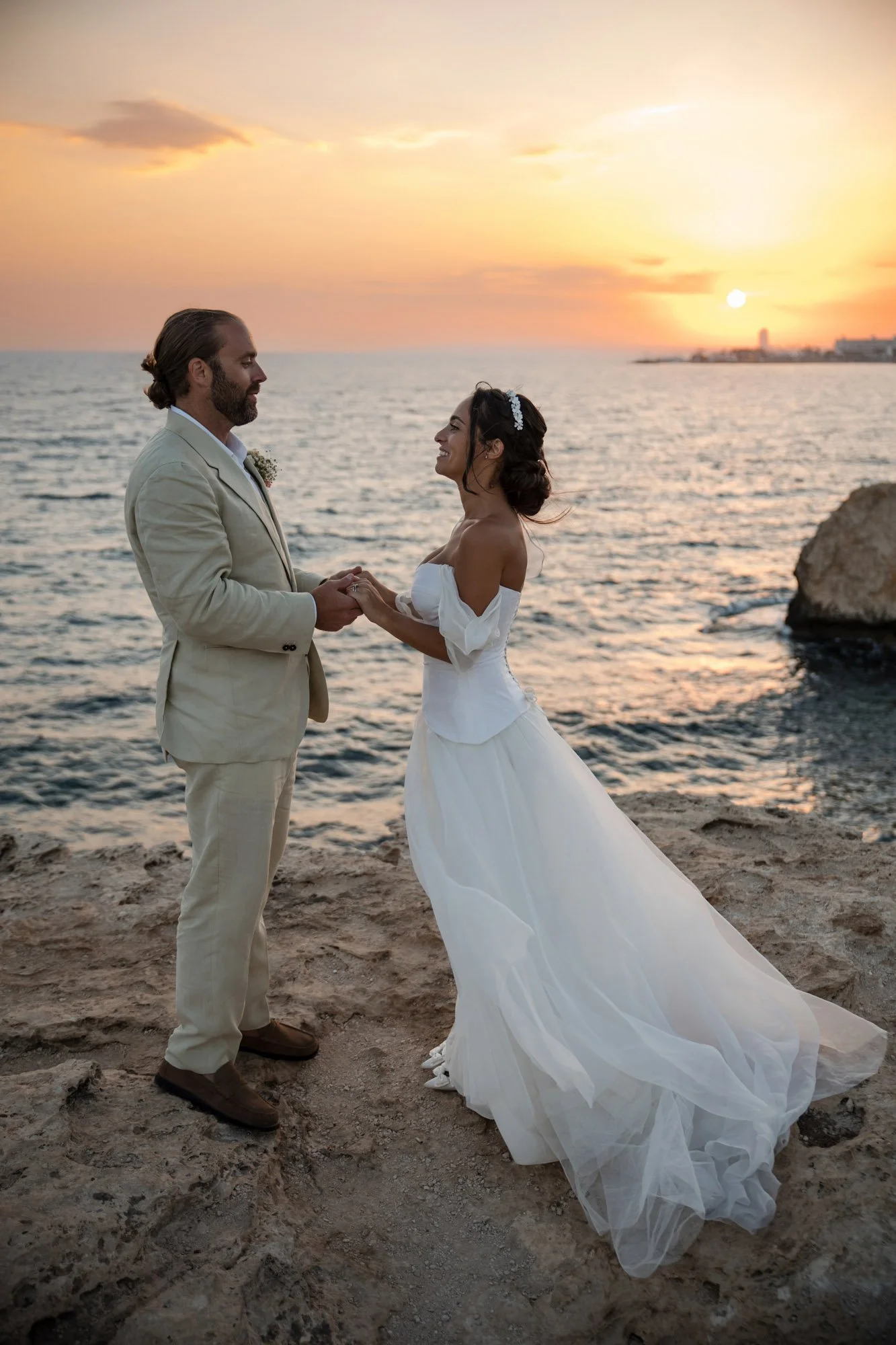 A bride and groom holding hands on a sandy beach at sunset, with the ocean and a city skyline in the background.