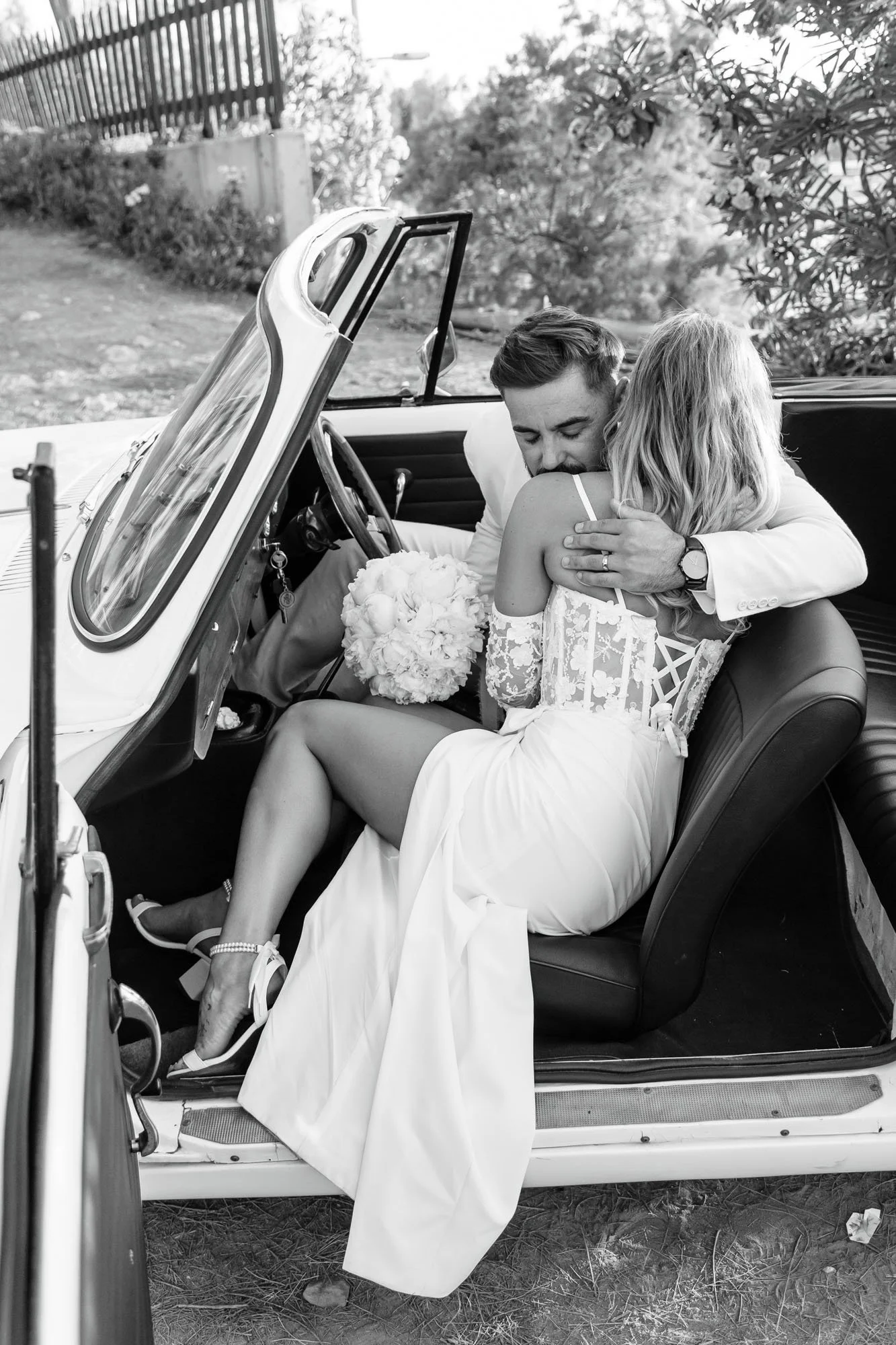 A black and white photo of a bride and groom embracing inside a vintage car, with the bride holding a bouquet of flowers.