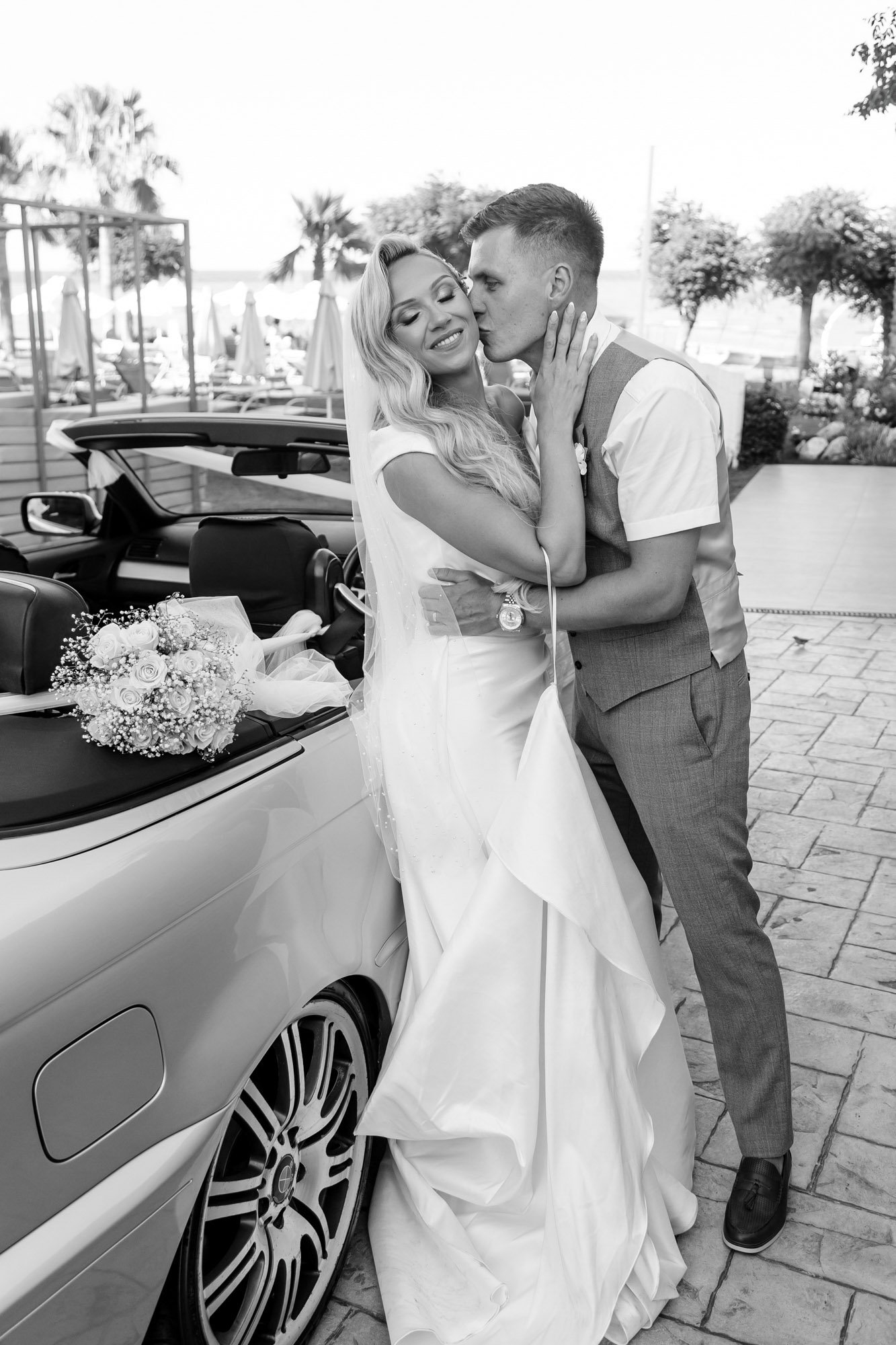 A newlywed couple sharing a kiss and embracing beside a convertible car decorated with a bridal bouquet, outdoors at a wedding or reception venue.