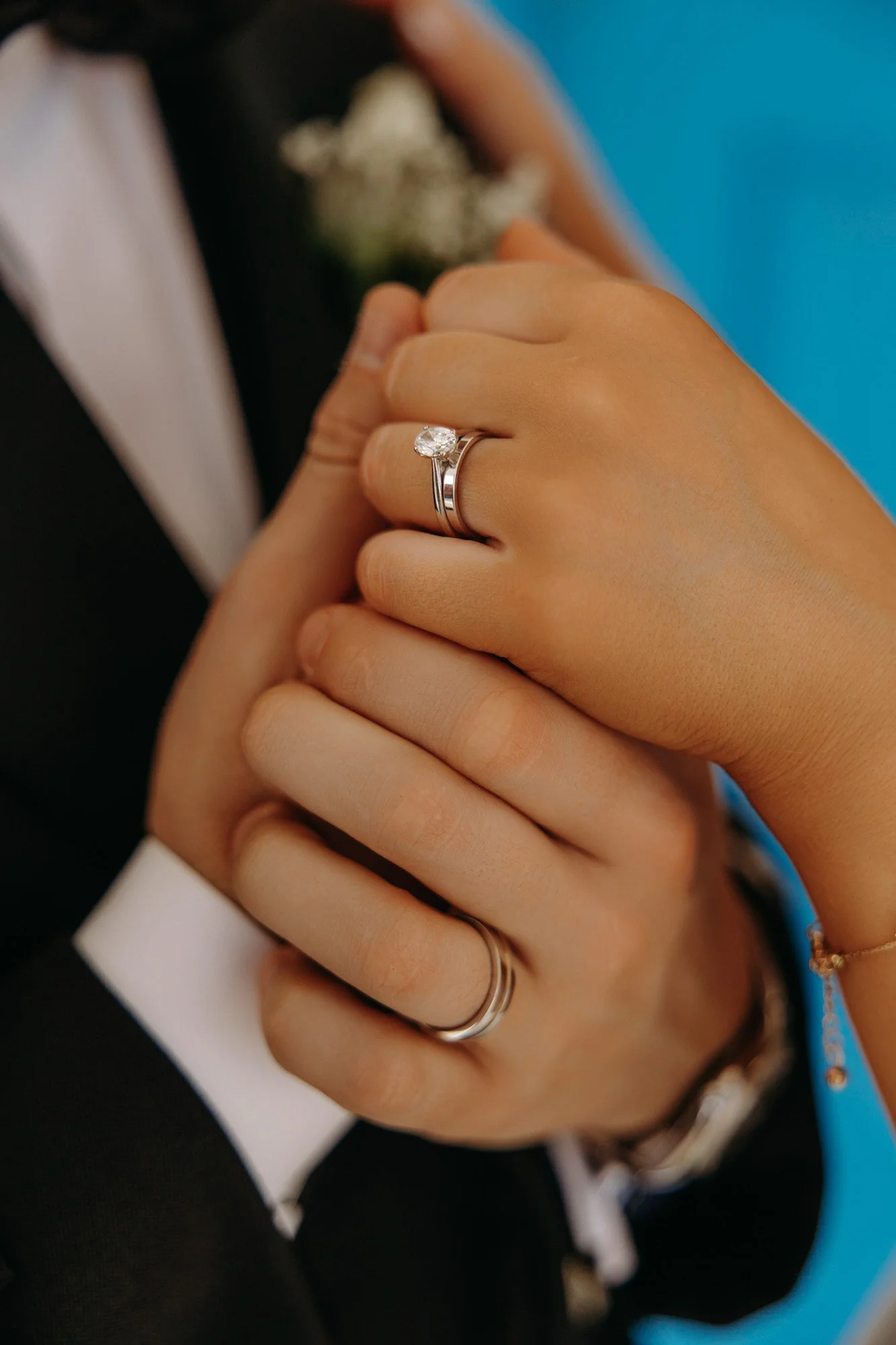 Close-up of a bride and groom holding hands, showing wedding rings; the bride's ring features a large diamond, and both are wearing simple silver bands. The groom is in a tuxedo, and the bride has a gold bracelet.