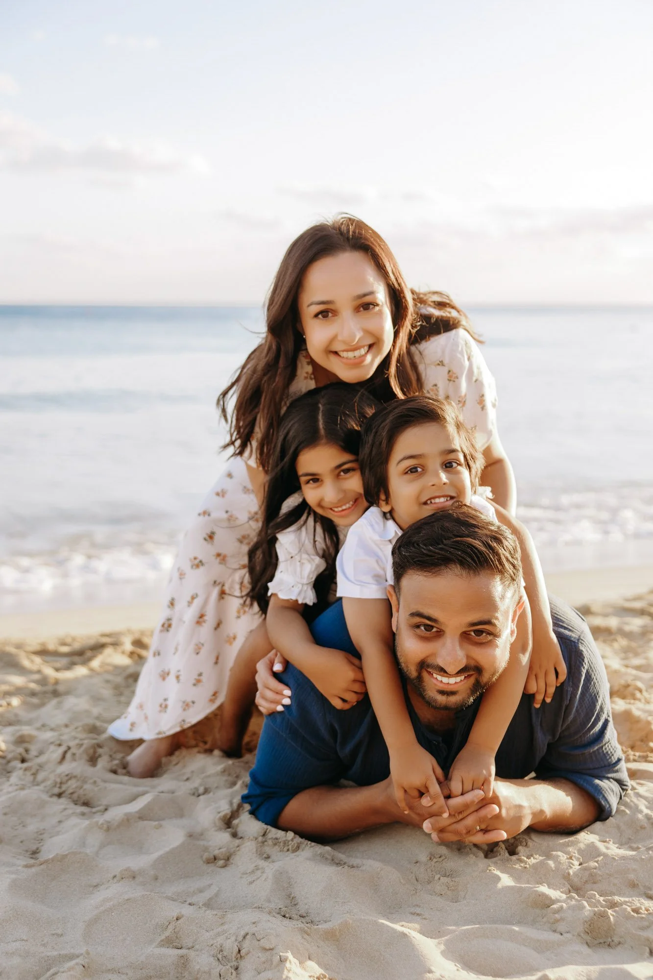 A family of five sitting and lying on the sandy beach, with the ocean and sky in the background, enjoying a sunny day.