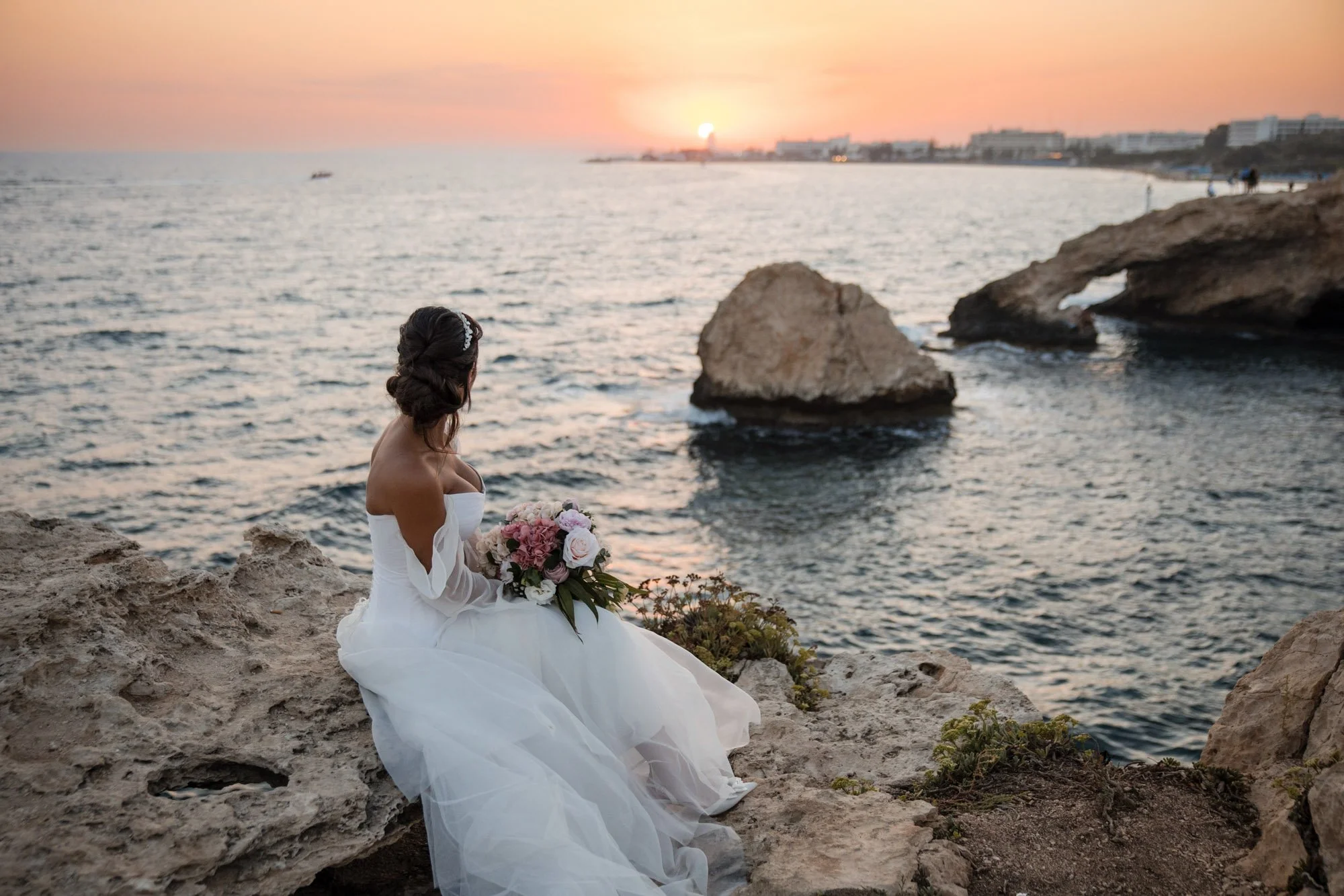 A bride in a white wedding dress sitting on rocky coastline holding a bouquet of pink and white flowers, sunset over the ocean in the background.