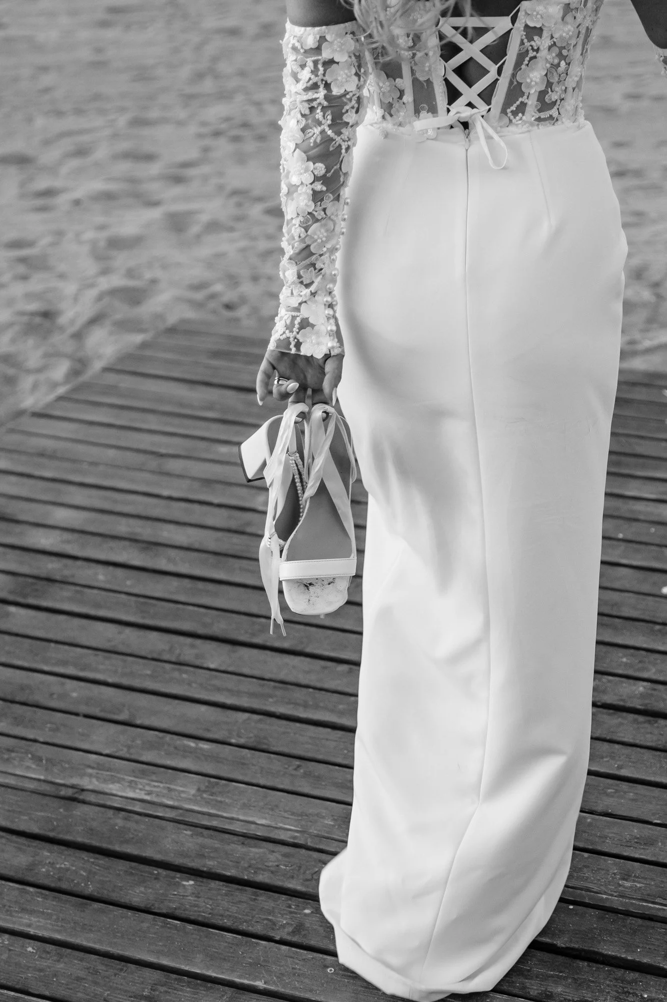 A person in a wedding dress holding a pair of high-heeled shoes with ribbons, standing on a wooden dock by the water.