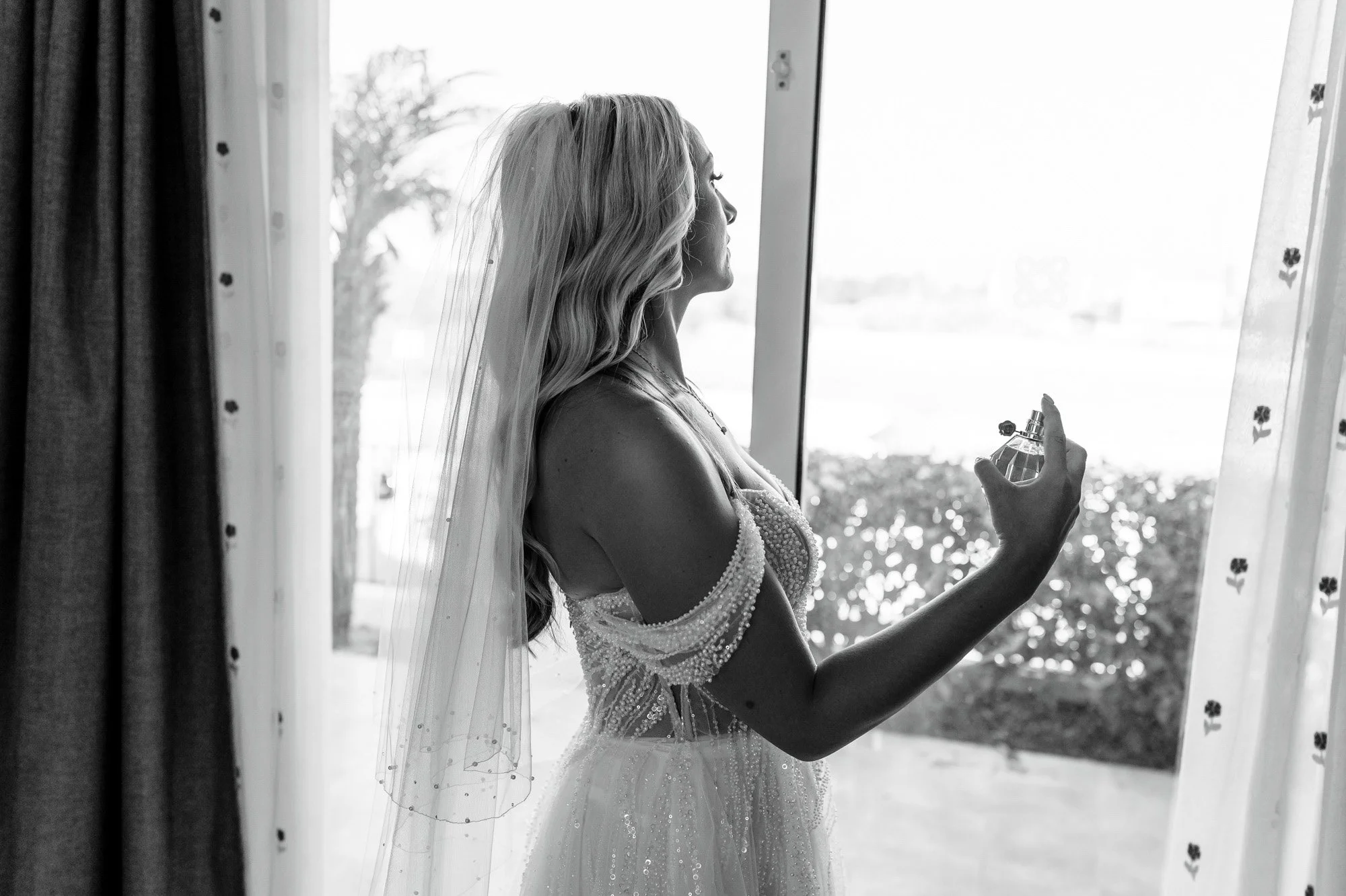 A bride standing by a window, holding a perfume bottle, dressed in a lace wedding gown with a veil, looking out.